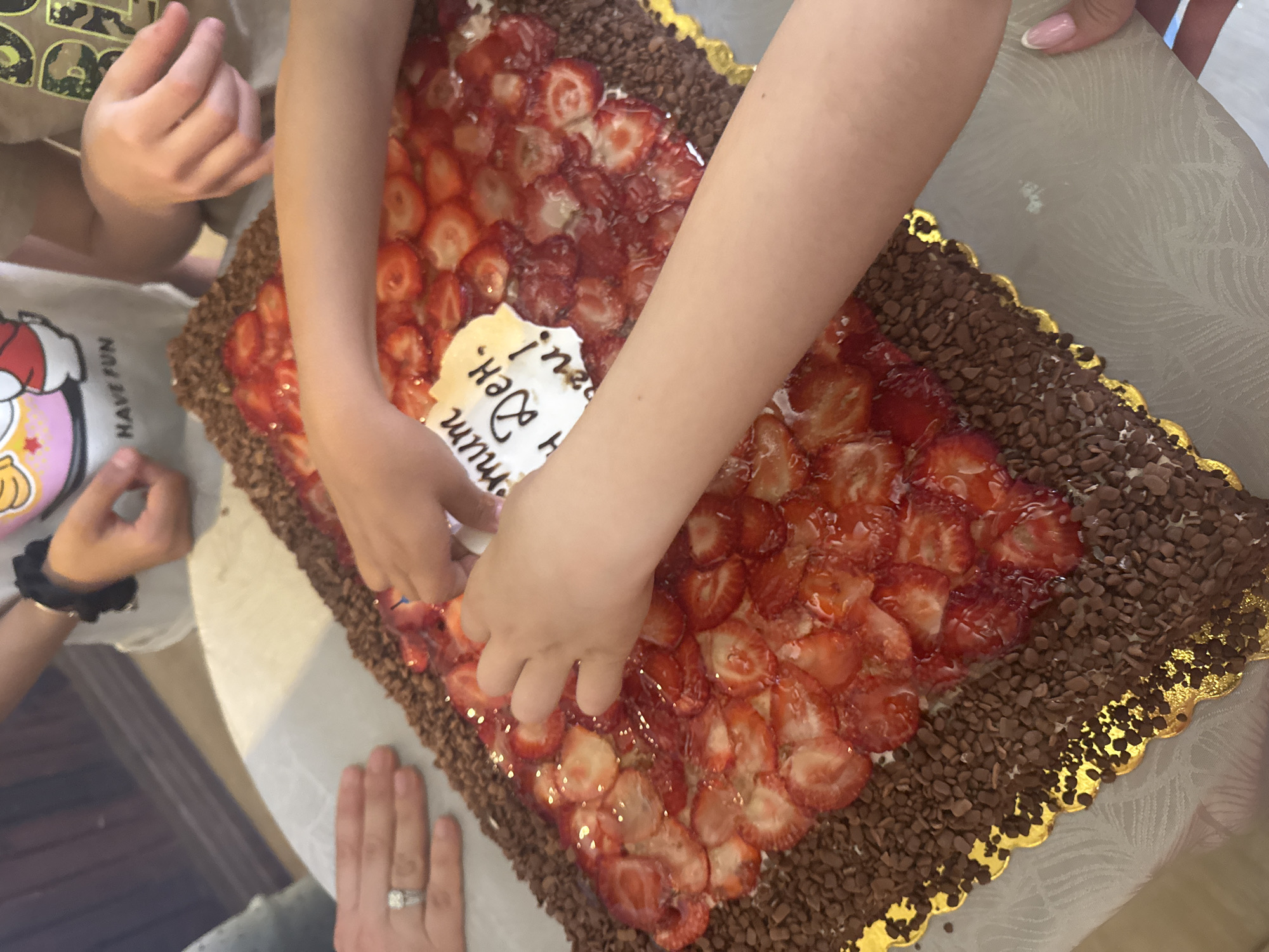 A top-down shot depicts a rectangular cake, likely a birthday cake, decorated with rows of sliced strawberries and chocolate sprinkles. A small white sign with Cyrillic text is placed in the center of the cake. Multiple hands reach towards the cake, indicating people interacting with it, presumably taking a piece. The surrounding environment is a table covered with a white patterned tablecloth. No specific details confirm the location is Elin Pelin, Bulgaria.