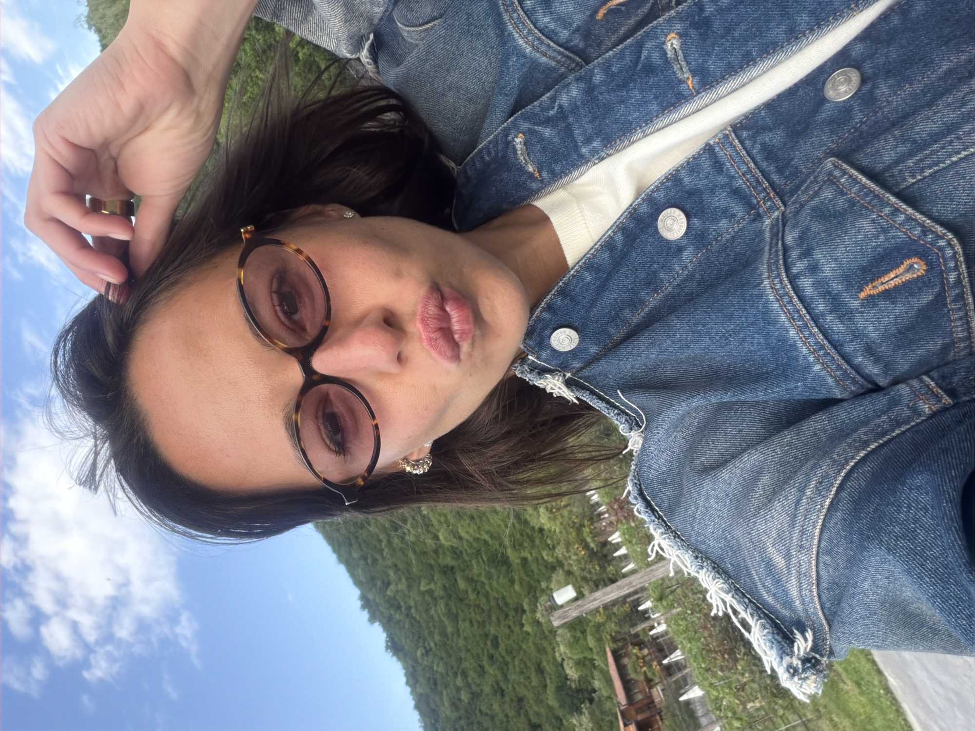 A woman is lying on her back outside in Napareuli, Georgia. She is wearing glasses and a denim jacket over a white shirt. Her right hand is touching her forehead. There are hills and a blue sky with clouds visible. There is also a vineyard.
