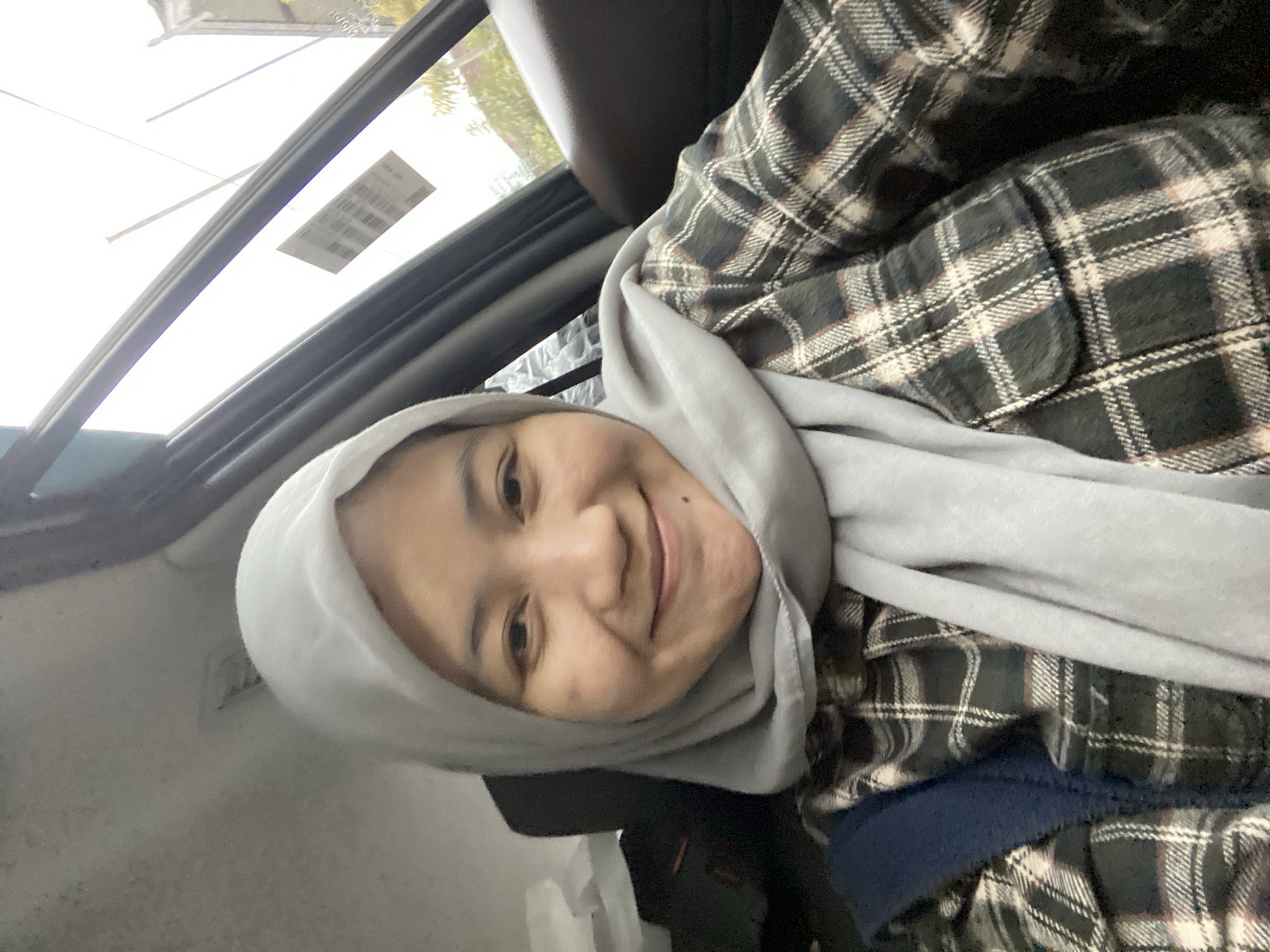 A woman wearing a light gray hijab and a plaid shirt is inside a vehicle. She is smiling and looking at the camera. The vehicle appears to be parked, and the woman is in the passenger seat. The background shows a window with some greenery and a sticker on the glass. The lighting suggests it is daytime. The scene is likely in Banjarbaru, Indonesia, given the context of the prompt.