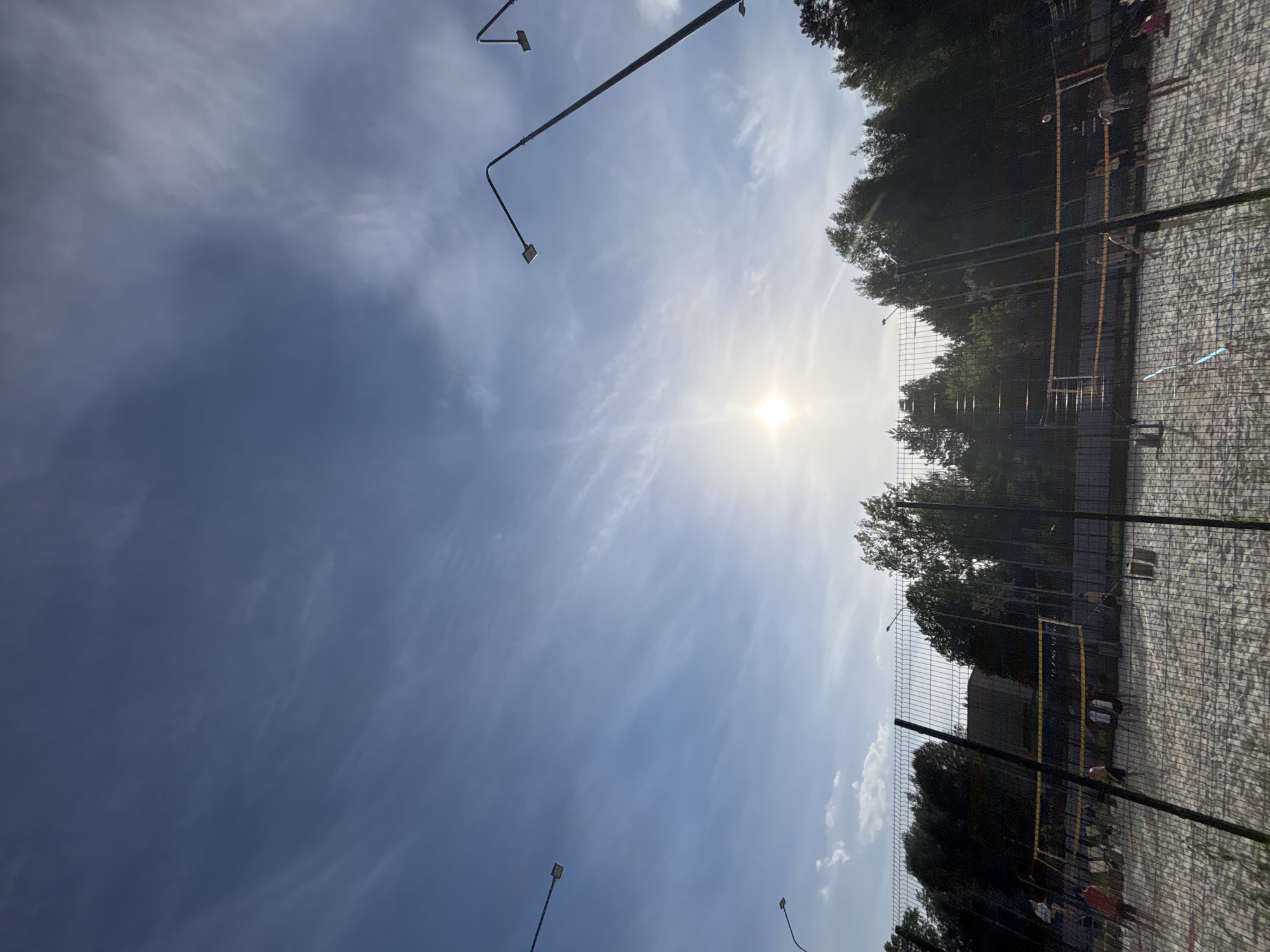 A vertical, eye-level shot presents an outdoor volleyball court under a bright, sunny sky. The sky is a mixture of light and dark blue hues, with scattered white clouds and the sun shining brightly. Trees line the periphery of the court, their dark green leaves contrasting against the lighter sky. Metal lampposts are positioned around the court, their sleek design adding a modern touch. The court itself is enclosed by a chain-link fence, visible in the foreground. Through the fence, figures of people can be seen, hinting at the presence of players or spectators. The ground of the court is covered in light-colored sand, characteristic of a beach volleyball setting. The environment suggests a city location, though the specific city and region are undetermined.