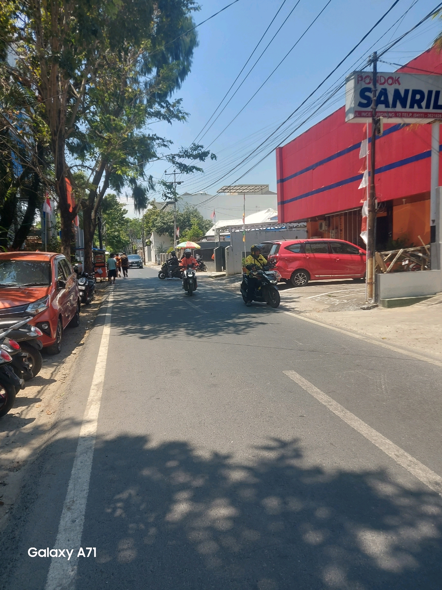 In Makassar, Indonesia, a street scene unfolds under a clear blue sky. The road, marked with a white line, is partially shaded by dappled sunlight filtering through trees on the left. Parked along the roadside are several motorcycles and an orange car. Further down the street, a red minivan is visible, parked in front of a bright red building with blue stripes. A sign above the building reads "Pondok Sanril". Several individuals are present; one person rides a black motorcycle in the foreground, wearing a yellow jacket and helmet. In the background, other figures are seen walking and riding motorcycles. The environment suggests a commercial or residential area with a mix of trees, buildings, and vehicle traffic.