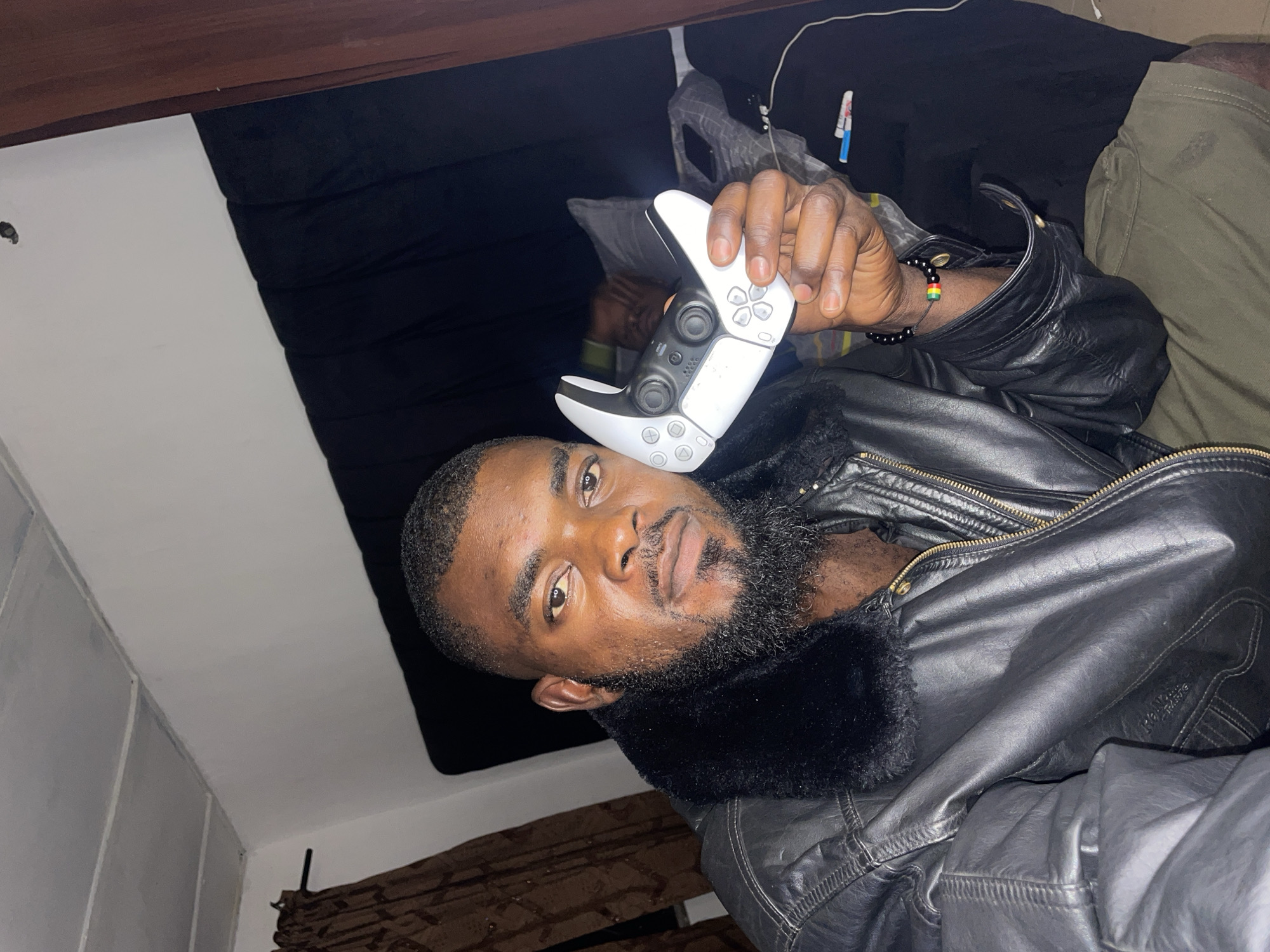 A bearded, brown-skinned man wearing a black leather jacket with a fur collar is holding a white PlayStation 5 controller. He is wearing a beaded bracelet on his left wrist. He is in a dark room, likely a bedroom. A black curtain hangs in the background, and a wooden structure is visible above the curtain. Additional clothing is visible. The location cannot be verified with certainty, only that he is purportedly in Jos, Nigeria.