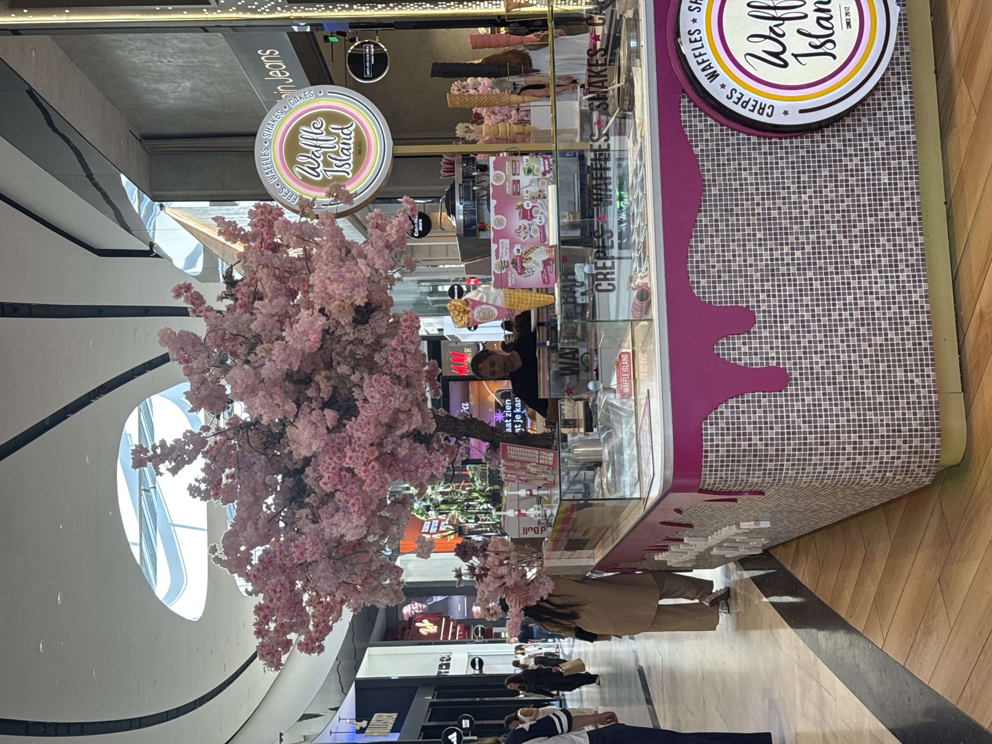 In the city of Leidschendam, Netherlands, within a modern shopping mall, a brightly decorated ice cream and waffle stand, "Waffle Island," is the focal point. A large artificial cherry blossom tree with delicate pink flowers is positioned to the left of the stand, adding a whimsical touch. A woman wearing a beige outfit stands behind the counter, appearing to be interacting with a customer. The stand features a display case filled with ice cream tubs and an assortment of waffle cones. Promotional posters showcasing menu items are visible on the stand. The surrounding environment includes shops with prominent signage, such as "Calvin Klein Jeans" and "Hollister." Other shoppers can be seen in the background, navigating the mall's polished floors and walkways. The scene captures a vibrant commercial setting within an indoor shopping complex.