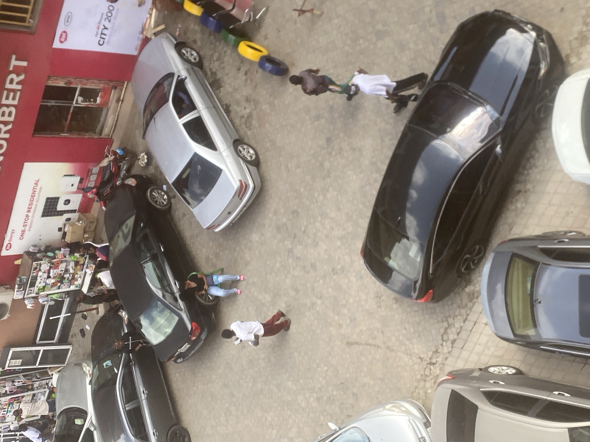 An aerial view reveals a street scene in Abuja, Nigeria, densely packed with cars parked on the street. In front of a building marked "NORBERT," a small market or vendor stall displays numerous items for sale. Several people are present: one person appears to be lying on the ground next to a car, another standing nearby, and two others are positioned near the sidewalk. Additional people can be seen around the vendor stalls. There are a few tires in blue, yellow and green sitting on the sidewalk.