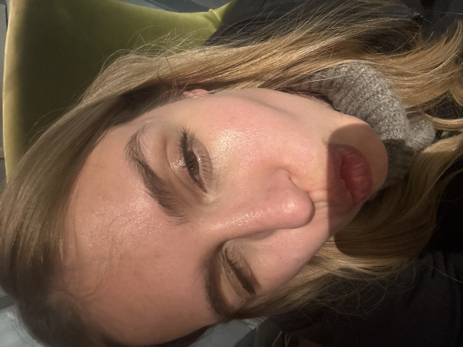 The image presents a close-up, eye-level selfie of a fair-skinned female individual. Her light brown to blonde hair is visible, styled with a slight part and framing her face. She is performing an action, with her right eye winking closed and her left eye open, appearing focused towards the camera. Her lips are pursed in a pouting or kissing gesture, partially obscured by a diagonal shadow cast across the lower half of her face, extending from her nose to her mouth area. She is dressed in a grey ribbed turtleneck sweater, worn beneath a dark outer garment. A small, thin silver hoop earring is visible in her left ear. The background is softly out of focus, revealing a plush, olive-green upholstered object, consistent with a chair or sofa, positioned behind her head to the upper right. The lighting is bright, suggesting a natural light source from above and to the right, illuminating her face and casting the shadow. The specific location within the city of Tbilisi, Georgia, cannot be determined from the visual information presented in the image.
