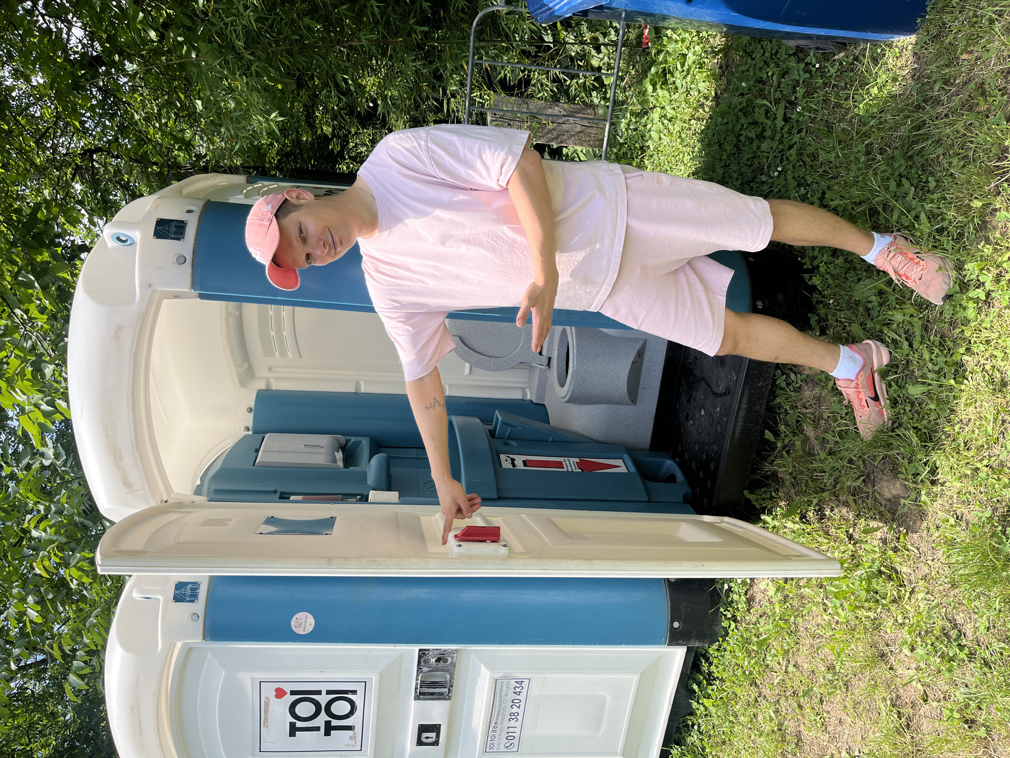 A young man stands outdoors next to an open blue and white portable toilet on a bright, sunny day. He is fair-skinned, wearing a light pink t-shirt, matching pink shorts, a pink baseball cap, white socks, and pink sneakers. He has a small tattoo on his left forearm. The man is smiling and looking directly at the camera, posing with his left hand gesturing towards the portable toilet's open door latch, and his right hand slightly extended.
The scene is set in a grassy area with lush green trees and foliage visible in the background, suggesting an outdoor event or recreational space. The bright sunlight casts clear shadows, indicating it is daytime. The portable toilet, an object often used at outdoor gatherings or construction sites, is a prominent feature.
Visible text on the side of the portable toilet includes the company name "ΤΟΙ ΤΟΙ d.o.o. Mobilni Sanitarni Sistemi" and an address "Bulevar Kralja Aleksandra 177, Beograd," along with a phone number "011 38 20 434." This text suggests the portable toilet belongs to a Serbian company, aligning with the provided location context of Sremski Karlovci Municipality, Serbia. The overall mood is casual and cheerful, with the man's pleasant expression contributing to the relaxed atmosphere.