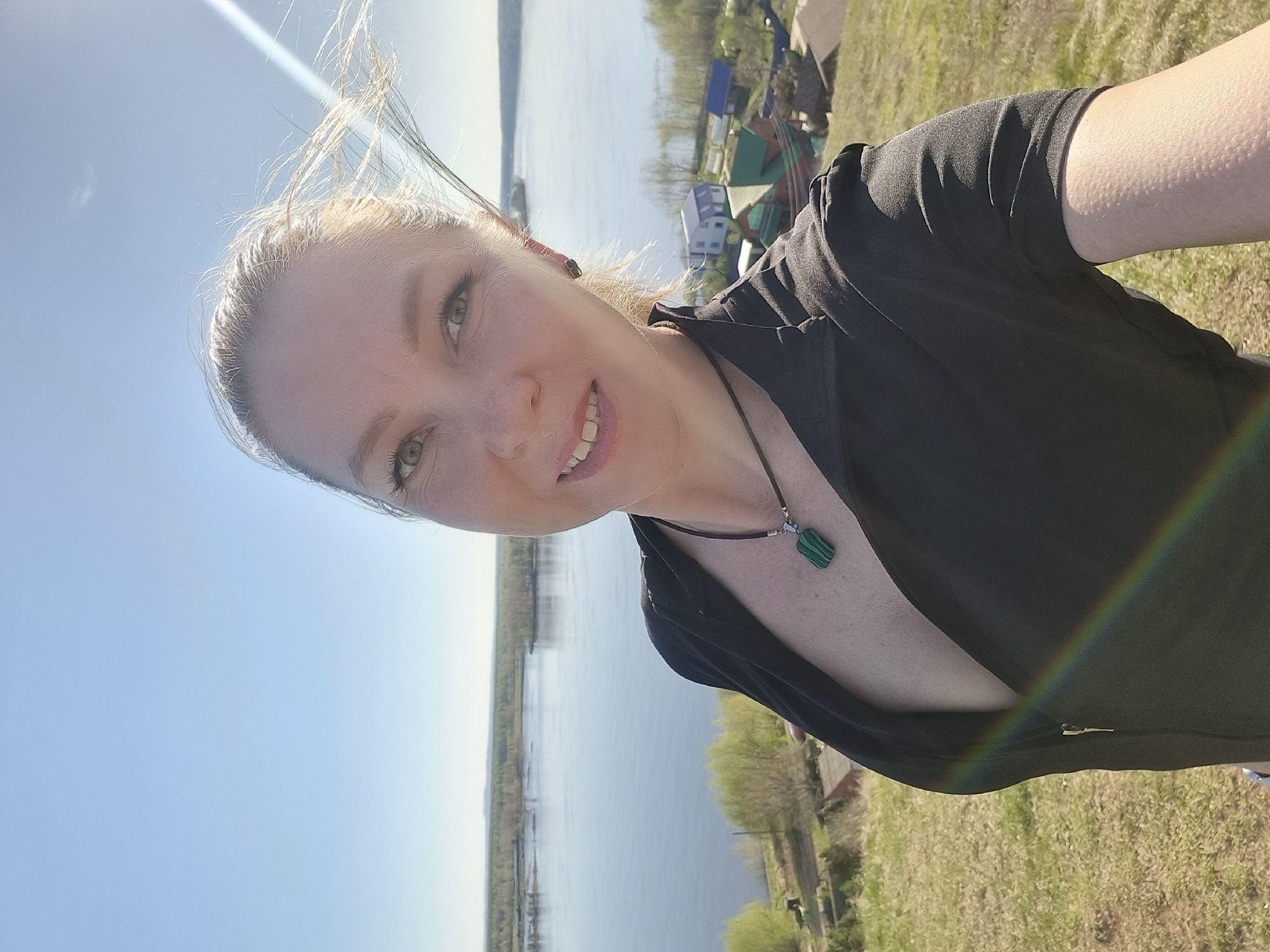 A selfie of a blonde woman is oriented at an angle, with the woman facing the camera and smiling. She is wearing a black top and a necklace with a green pendant.
The background consists of a landscape featuring a body of water, likely a river or lake, with a shoreline visible on the opposite side. There are green trees and grass, suggesting a natural or park-like setting. In the upper right corner, there are rooftops of buildings that could be part of a small town or village. The sky is clear and blue.
The location is Ольховка, Russia.