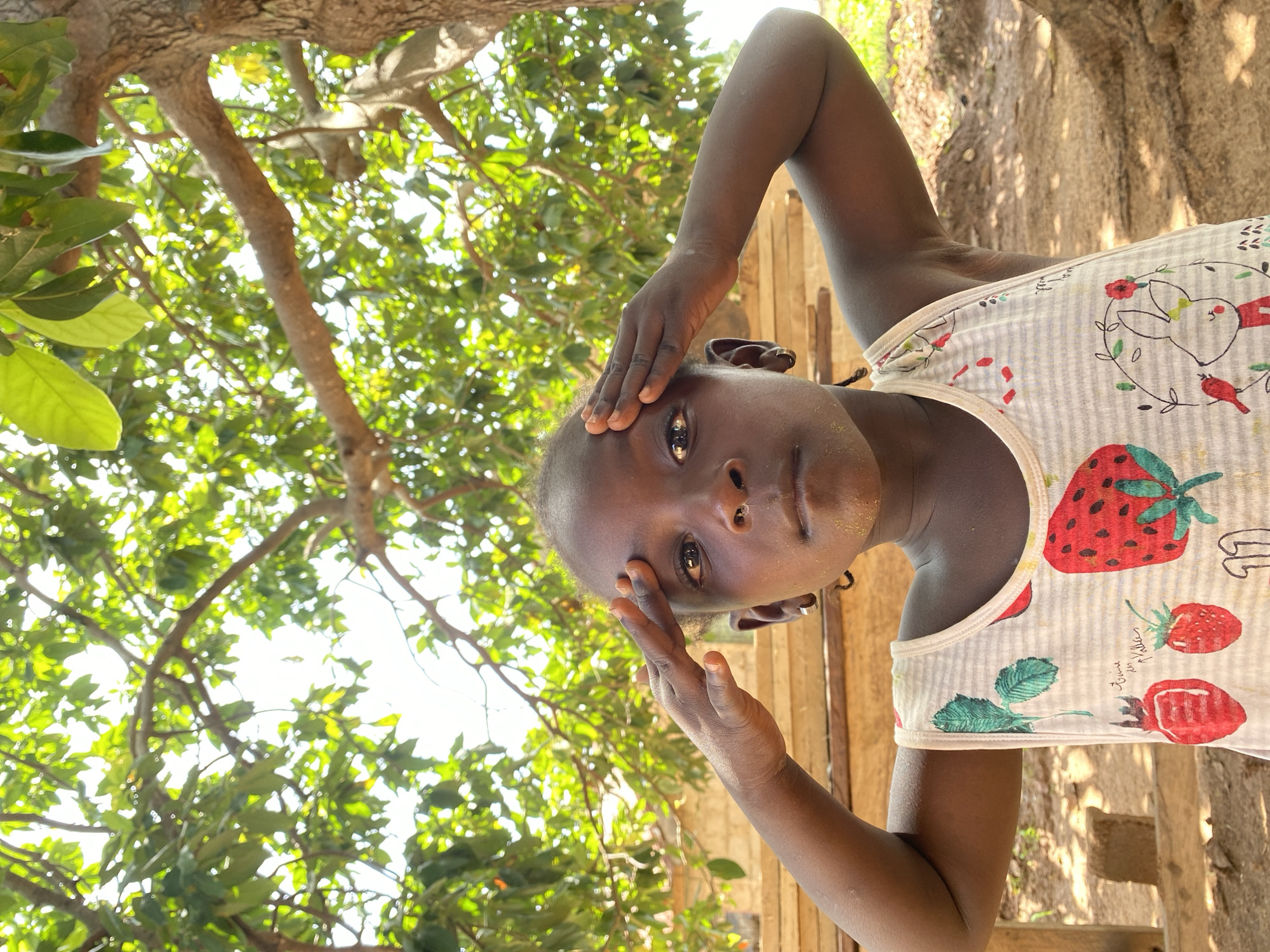 A young person with brown skin stands outside in Riyom, Nigeria. The person is positioned in front of a tree with green leaves. The young person is wearing a white tank top with a print of strawberries, rabbits, and birds. The person has their hand on their forehead. The sky is visible through the tree branches. The environment is natural and sunny.
