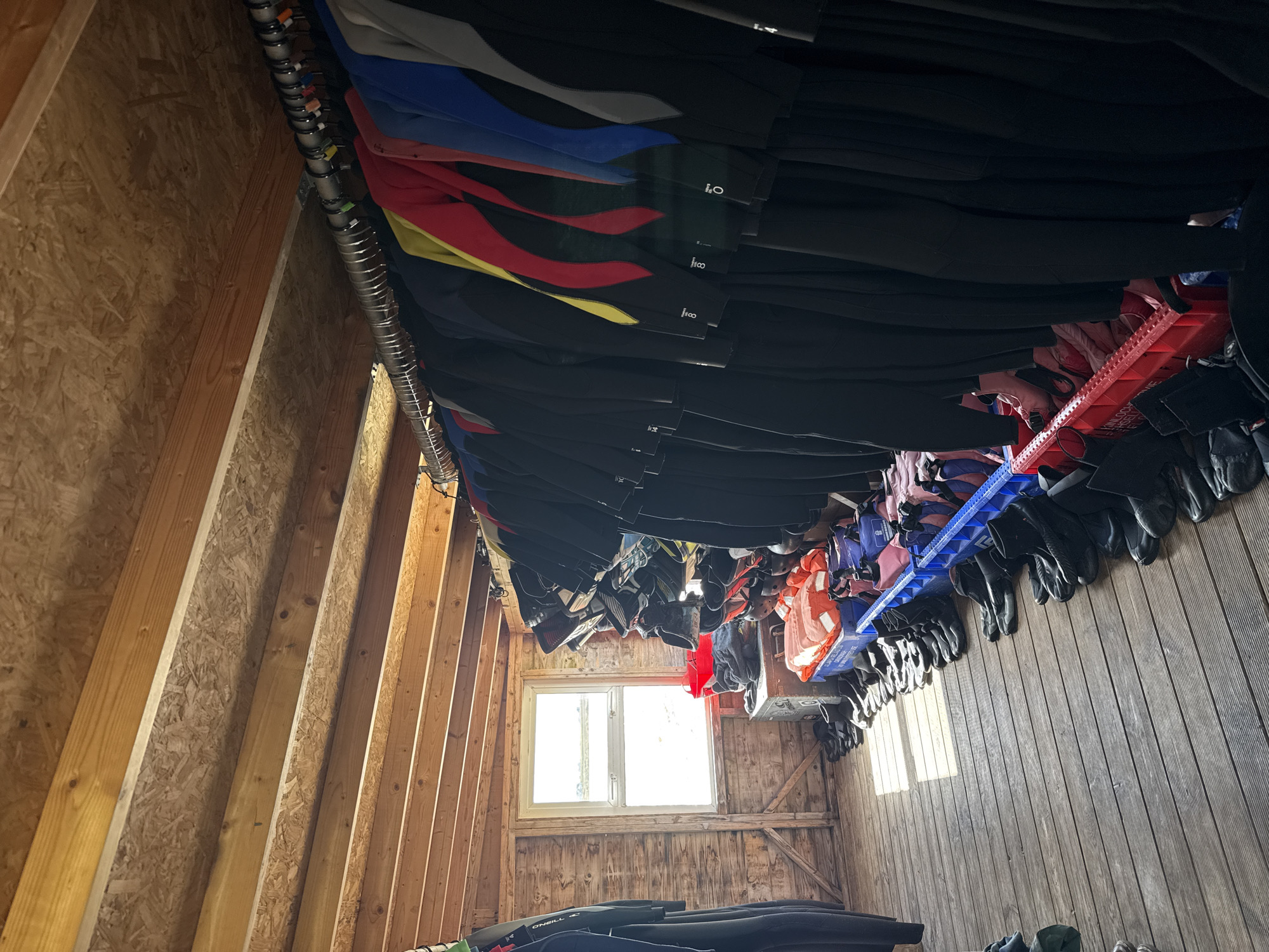 The image shows the interior of a shed-like structure in an unknown location. The walls are made of wood paneling and exposed wooden beams. A window provides natural light. Wetsuits of varying colors and sizes hang from a metal rack, and life jackets are placed in red and blue plastic storage crates. Gloves and possibly other aquatic gear are stored on the walls. Some black wet suits are laid on the floor.