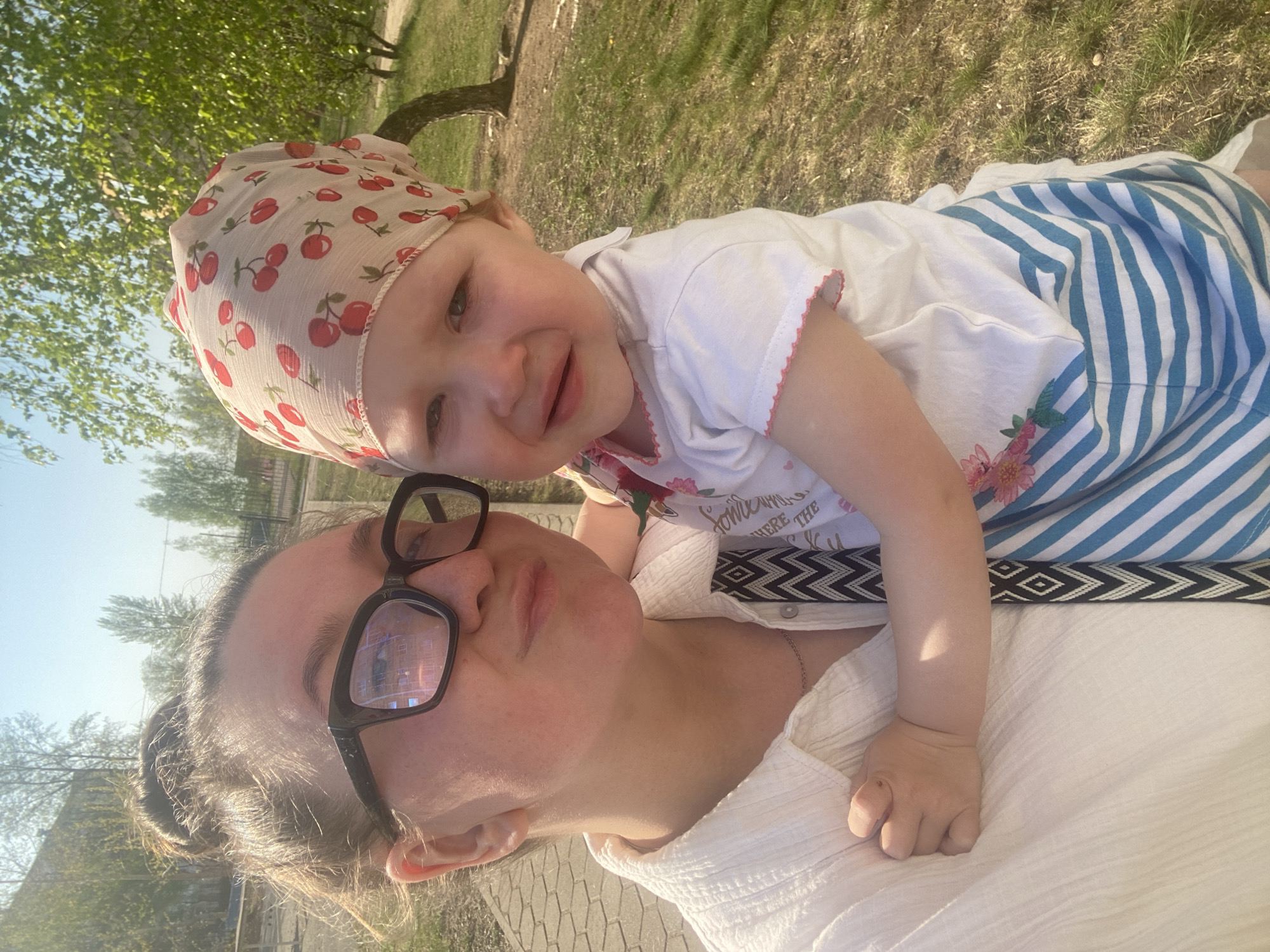 The image shows a woman with glasses and a young child in an outdoor setting. The child is wearing a cherry-patterned headscarf and a dress with blue and white stripes. The woman is wearing a white shirt and appears to be on a sidewalk or paved area, with a grassy area and trees in the background. The photo was taken in the city of Chaikovsky, Russia.