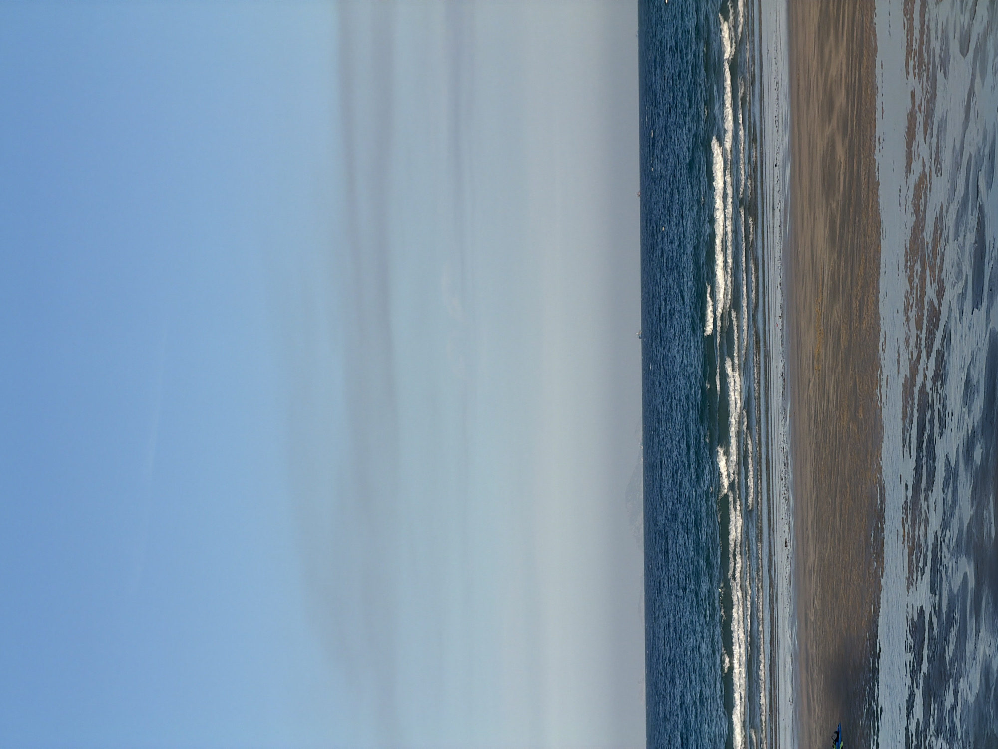 This is a vertical, medium-shot, eye-level photograph of a beach at Cam Lam, Vietnam. The image is divided into three horizontal sections: sky, sea, and shore. The sky is a pale blue with faint horizontal cloud formations. The sea is a deep blue, with several lines of white waves crashing on the shore. The shore consists of wet sand with tidal pools reflecting the sky and sea. In the distance, on the horizon, are a few faint shapes that appear to be ships or islands. In the lower left corner, a small portion of a blue and green object, possibly a kite or beach toy, is visible. The overall scene is serene and captures the natural beauty of the coastline.