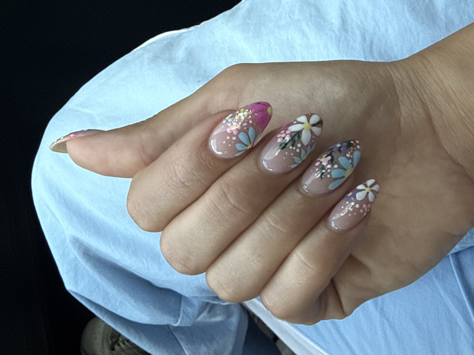 A close-up, vertical image depicts a person's left hand, exhibiting five elaborately decorated fingernails. The hand, displaying a light to medium skin tone, is positioned with fingers slightly raised and extended. The nails are almond-shaped with a clear or nude base coat. Each nail features intricate floral designs and scattered embellishments. Specific designs include white flowers with yellow centers, light blue flowers, pink and purple accents, and green leaves. The ring finger's nail additionally displays a vibrant pink tip with glitter and small blue and pink specks. The hand rests on or against a light blue fabric, possibly an article of clothing, which occupies the lower left portion of the frame and shows soft folds. The background to the upper right is dark and indistinct, suggesting a shadowed interior space. The scene is observed to be within the city of Macon, United States of America.
