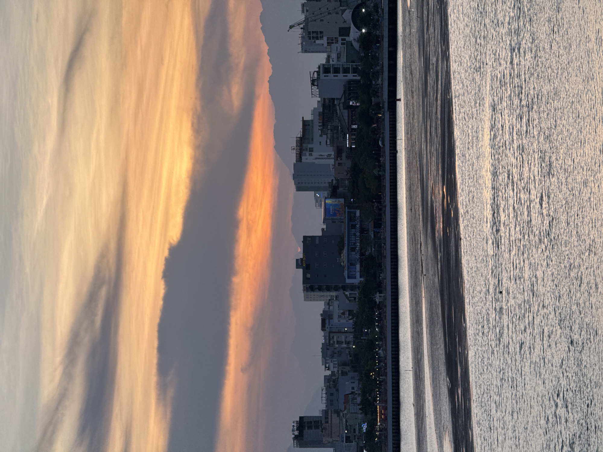 This image captures a sunset over the city of Da Nang, Vietnam. The sky is a vibrant display of orange, yellow, and gray clouds, with the warm tones of the sunset illuminating the horizon. In the foreground, the water of the Han River is calm, reflecting the colors of the sky. A large expanse of sand or mudflats is exposed along the riverbank, showing ripples and patterns from the receding tide.
Dominating the mid-ground is the Da Nang cityscape, a collection of modern buildings of varying heights, including high-rises and lower structures. Some buildings are illuminated, suggesting it is dusk. Behind the city, a range of mountains forms a silhouetted backdrop against the colorful sky. The overall scene depicts a serene and picturesque urban landscape at the end of the day.