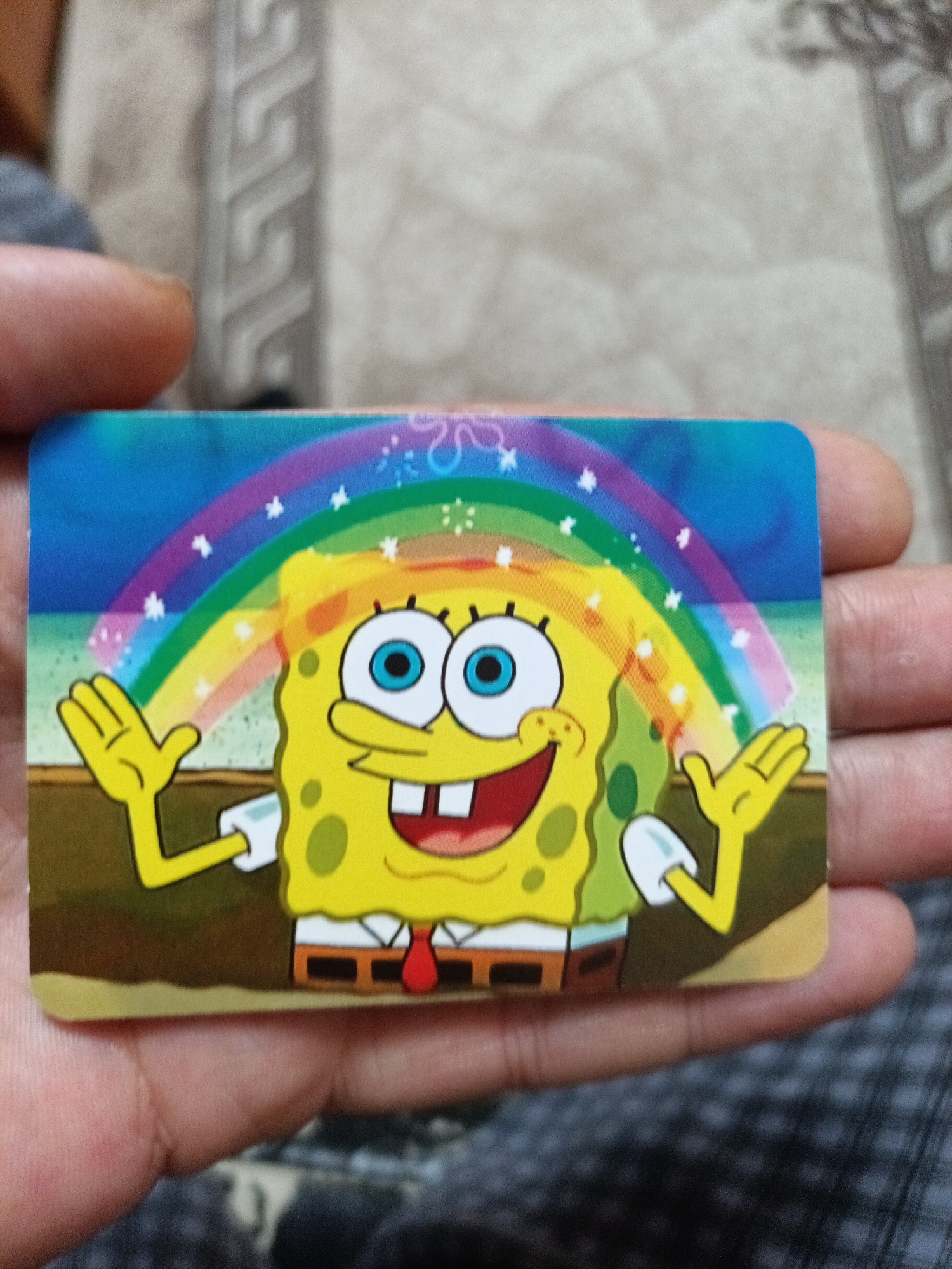 In the original image, a Spongebob Squarepants sticker is held by a person's hands. The sticker depicts Spongebob with a wide smile, blue eyes, and a red tie, standing in front of a rainbow. Small white stars are scattered around the rainbow. The background behind the sticker appears to be a textured surface with a repeating geometric pattern, possibly a blanket or a rug. The person's fingers are visible on the sides and bottom of the sticker, holding it flat. The image is taken from a slightly elevated angle, looking down at the sticker and hands. There is no discernible information in the image that would allow for the identification of the city of Nyagan, Russia, or any specific urban or natural environment.