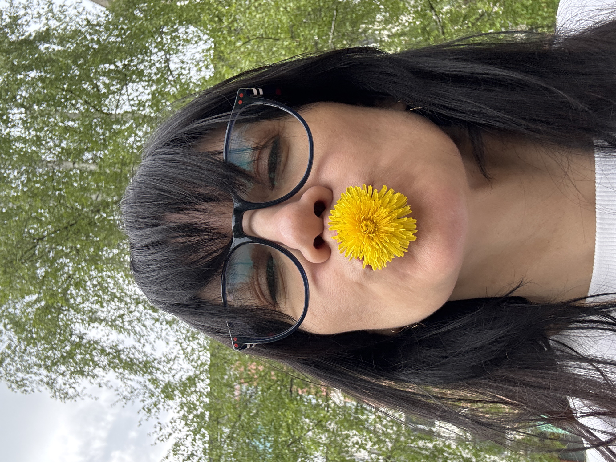 A woman with dark hair and glasses is posing with a dandelion in her nose. The background shows green trees, suggesting an outdoor setting. It is not possible to definitively determine the location is the city of Chaikovsky, Russia from this image alone.