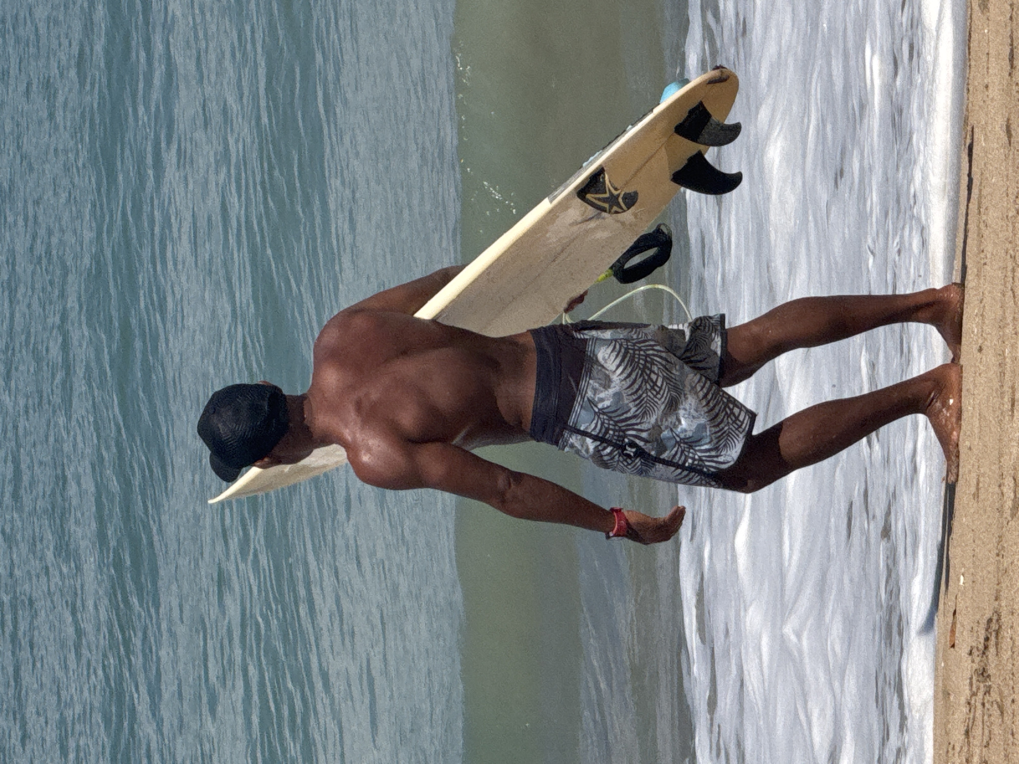 A shirtless man wearing a black baseball cap and patterned swim trunks is walking on a sandy beach towards the ocean. He is carrying a light-colored surfboard under his right arm. The surfboard has black fins and a leash. The ocean water is blue with white foamy waves reaching the shore. The sun is shining, casting a shadow of the man on the sand. The man is standing in the water, with his feet partially submerged. The scene appears to be a typical day at the beach, likely in a location known for surfing.