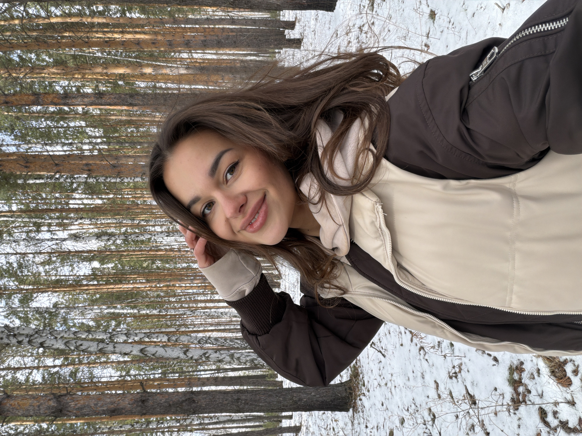 A young woman poses for a selfie in a snowy forest. She is wearing a brown and beige bomber jacket. Tall trees surround her, their trunks appearing to be birch and pine. The ground is covered in snow. The photograph likely depicts a natural area located near or in the city of Krasnoyarsk, Russia.