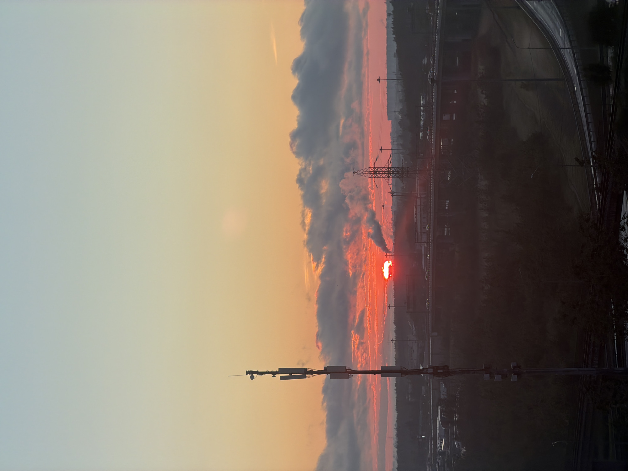 The image depicts an urban landscape at sunrise or sunset, likely on the outskirts of a city. The sky is a gradient of soft blues and oranges, with clouds tinged red by the low sun. In the foreground, a tall communication tower with multiple antennas is prominently featured on the left side. Beyond it, the scene opens up to a sprawling city view. Industrial structures, including smokestacks emitting plumes of smoke, are visible. A highway overpass with a few cars on it crosses the middle ground, and further in the distance, more buildings and infrastructure blend into the hazy horizon. The overall impression is one of an industrialized urban environment under a dramatic sky.