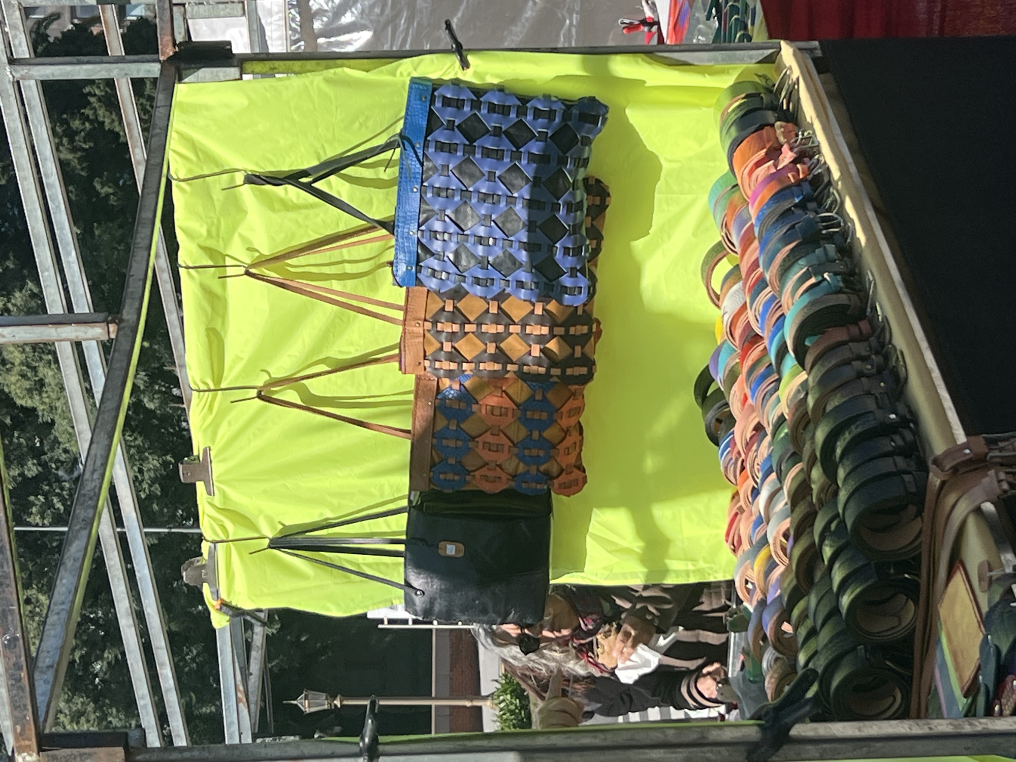 The image shows a street vendor's stall in Buenos Aires, Argentina. Displayed against a bright yellow backdrop are four handbags in various colors and textures. To the right, a vertical stack of colorful leather belts is arranged. A person, whose upper body is partially visible, is standing near the stall, possibly interacting with the merchandise. The stall is constructed from metal poles and beams, suggesting a temporary or semi-permanent structure. The backdrop is a lime green sheet or cloth. The handbags are hung in a vertical row and appear to be crafted from woven or braided materials. The belts vary in color, and their buckles are visible. The overall setting implies an outdoor market or fair environment.