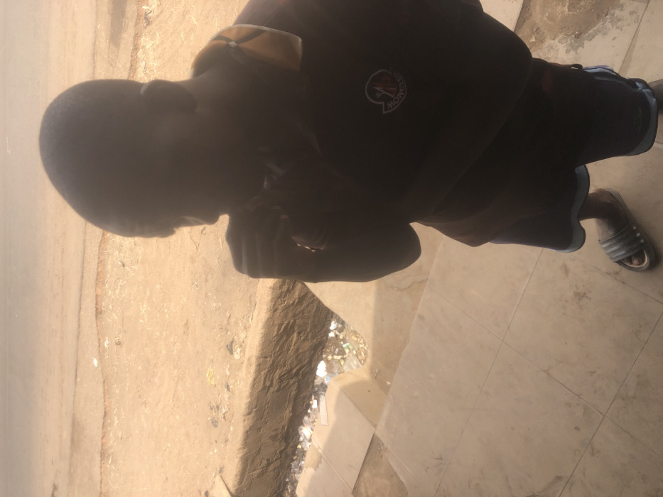 The image depicts an outdoor scene in Kufena, Nigeria, viewed from a slightly elevated perspective. A single male individual is positioned in the foreground, facing partially away from the camera and towards the left. His facial features are obscured by shadow, but his right hand is raised near his mouth or chin. He wears a dark short-sleeved polo shirt with a yellow-gold collar and a circular Moncler emblem on the left chest. Below, he wears dark shorts with a light blue stripe along the bottom edge, and light-colored slide sandals on his feet. The individual stands on a light-toned, tiled surface. Immediately beyond this surface, a low concrete curb defines the edge of a narrow ditch or gutter filled with miscellaneous refuse, including plastic containers and paper debris. Further in the background, a wide, unpaved, dusty road extends horizontally across the frame, exhibiting minor surface undulations. The scene is illuminated by bright outdoor light, casting the foreground subject into relative shadow.
