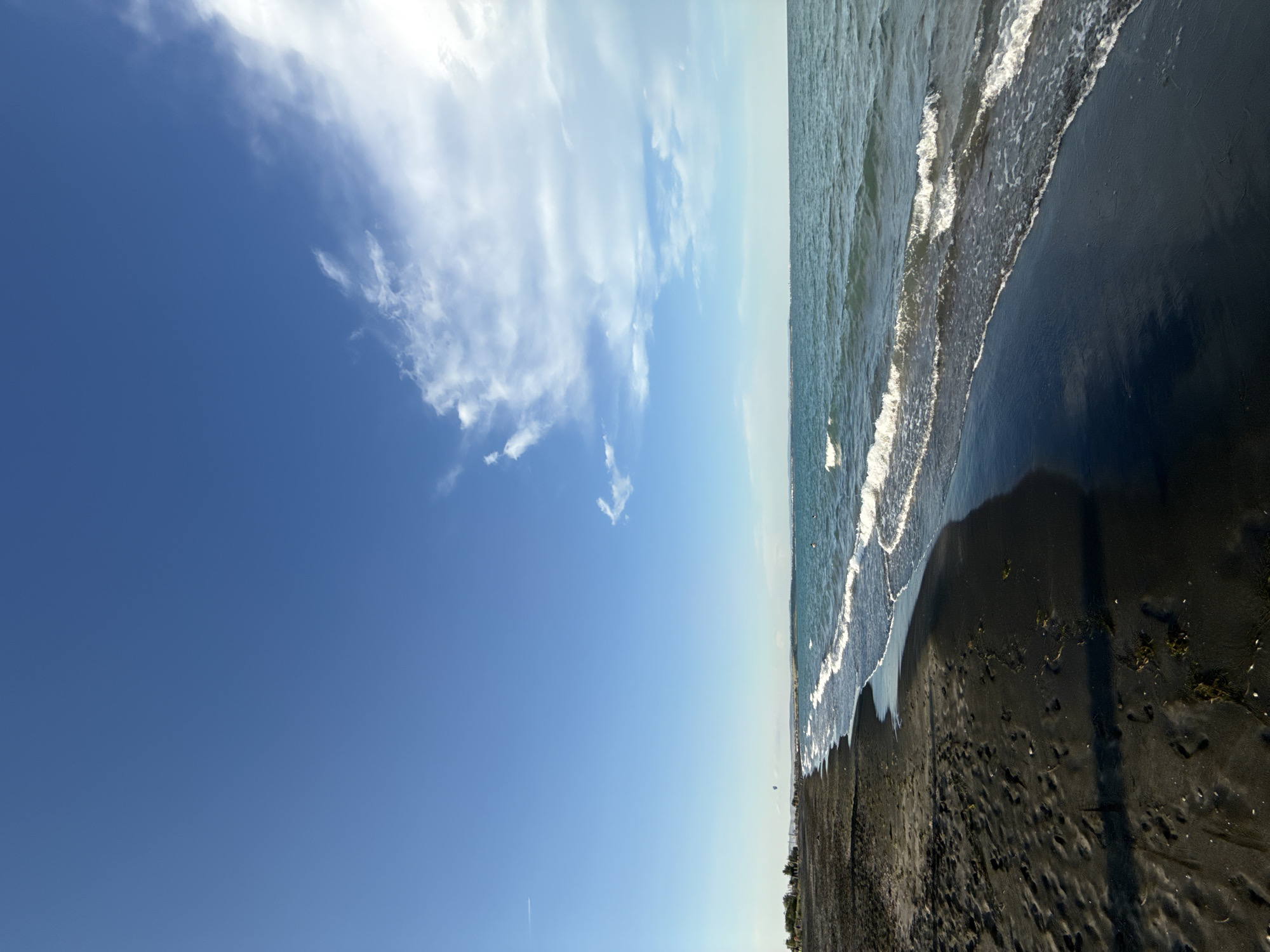 The image depicts a dark sandy beach with gentle waves lapping at the shore under a clear blue sky with scattered white clouds. In the distance, a thin strip of land with some trees and buildings is visible, suggesting a coastal settlement. A few kite surfers can be seen in the water, indicating recreational activity. The scene appears to be captured during daylight hours, possibly late afternoon due to the shadows on the sand. The location is identified as the beach in Burgas, Bulgaria, a coastal city known for its sandy beaches and proximity to the Black Sea.