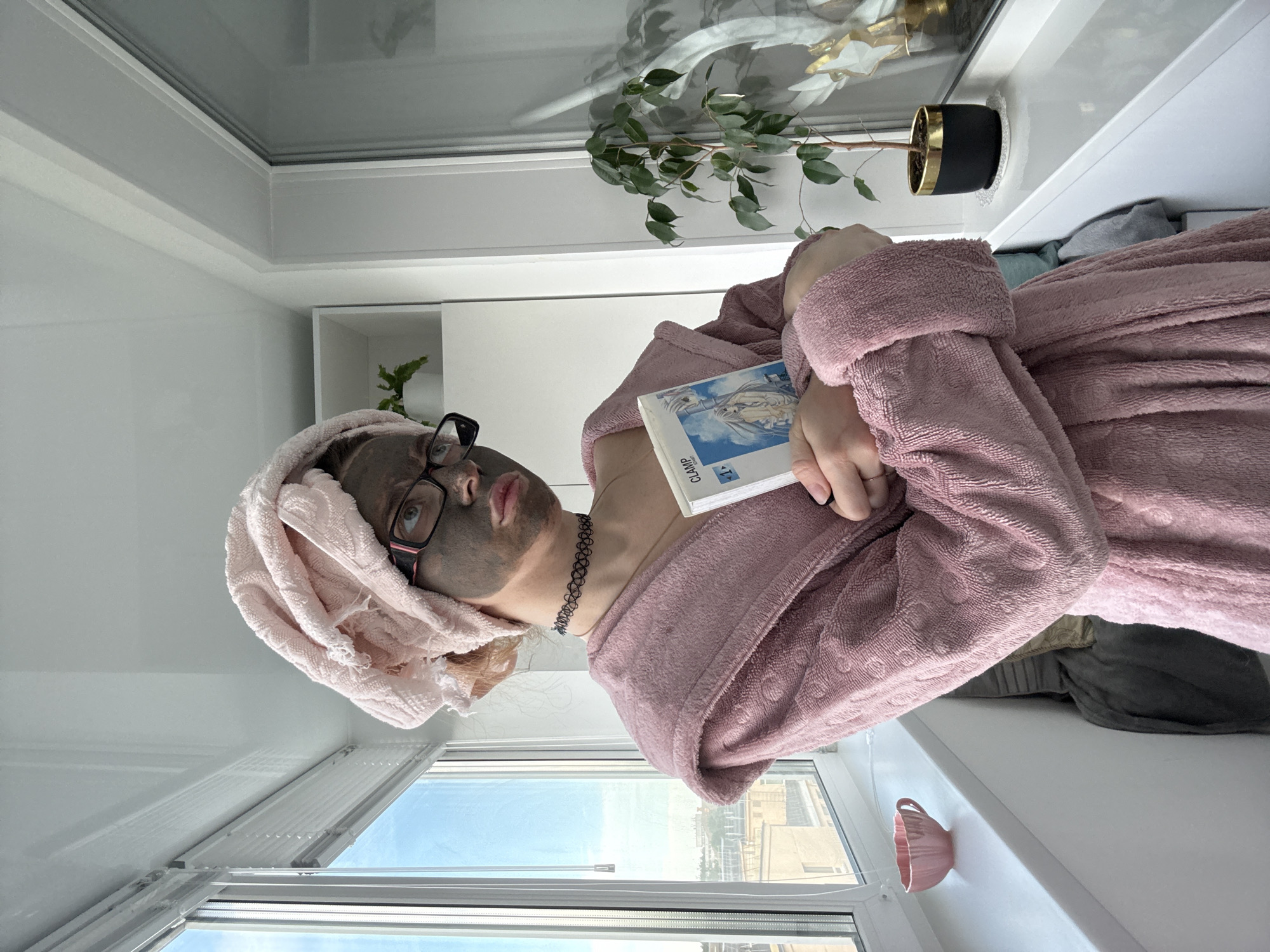 A young woman with a pink towel wrapped around her head and a clay mask on her face is standing on a balcony. She is wearing a pink bathrobe and glasses. She is holding a book titled "CLAMP" in her hands. A small pink teacup is on the windowsill to her left, and a potted plant is on the windowsill to her right. The balcony overlooks a city.