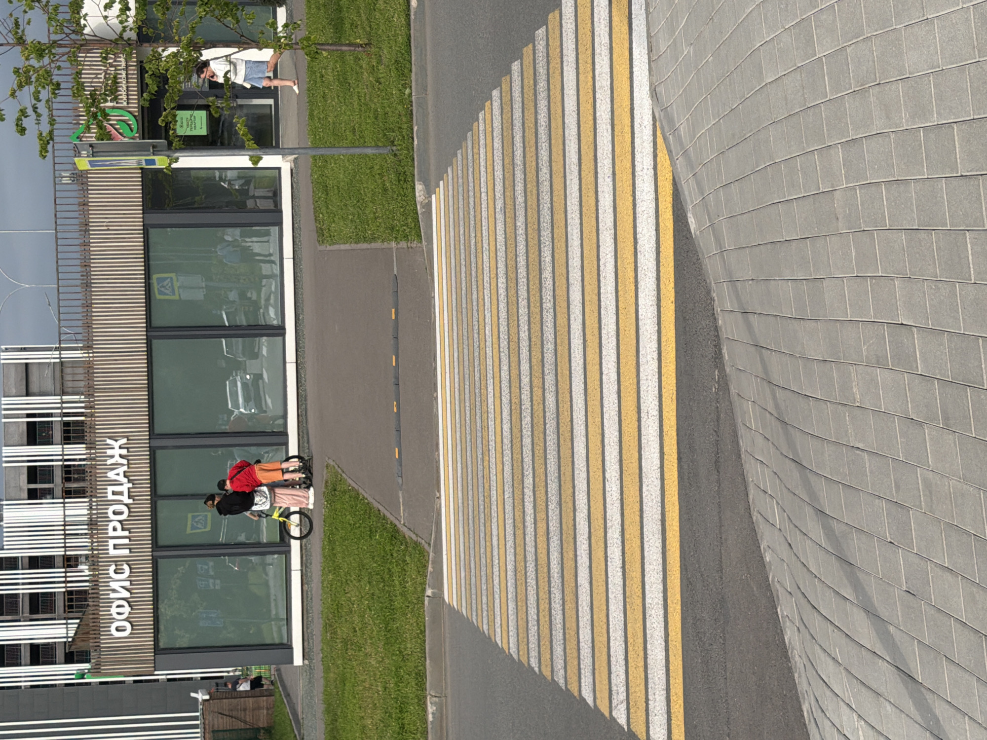 The image depicts an outdoor scene with a pedestrian crossing, a modern building, and a grassy area. Two people are visible near the building, one on a bicycle. The person in front is wearing a black hoodie and pink pants, and the person behind them is wearing a red jacket and orange shorts. They appear to be interacting with the bicycle.
The building has large glass windows and horizontal wooden paneling. Text is visible on the building's facade, reading "ОФИС ПРОДАЖ," which translates to "SALES OFFICE." A green sign with white text is also present on the building, but the text is not legible. A yellow and white striped pedestrian crossing is prominently featured in the foreground, suggesting a public area. The surrounding area includes a well-maintained lawn and a sidewalk paved with gray rectangular tiles. The lighting suggests it is daytime, and the weather appears to be clear. The scene implies an urban or suburban environment.