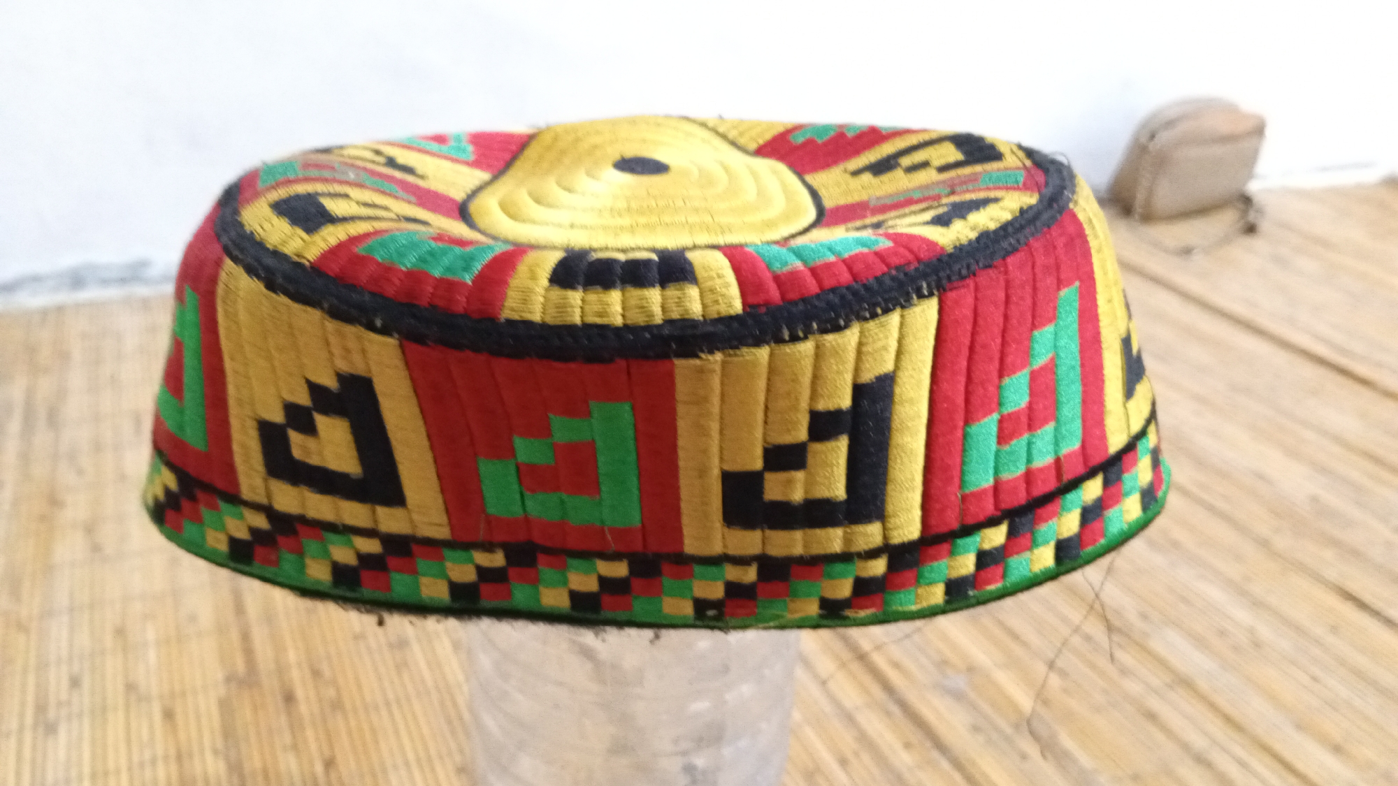 The image shows a traditional Indonesian hat, possibly a peci or songkok, resting on a clear plastic container. The hat is cylindrical with a flat top and a colorful embroidered pattern consisting of red, yellow, green, and black geometric shapes. The top of the hat is predominantly yellow with a concentric design and a small dark spot at the center. A black band separates the top section from the sides. The lower edge of the hat features a checkered pattern. The background includes a light-colored wall and a bamboo-like matting surface. A blurred tan object resembling a small bag or pouch is also visible in the background. The image was likely taken indoors. While the context specifies Lhokseumawe, Indonesia, the image itself doesn't provide visual clues to confirm the location.