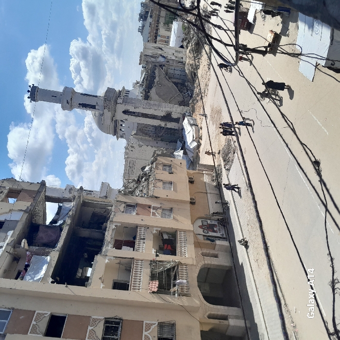 The image portrays a scene of extensive urban destruction in Khan Yunis, Palestinian Territory, under a partly cloudy blue sky. A wide, unpaved street dominates the foreground, with numerous dark overhead utility lines creating shadows. To the left, a multi-story, light-colored building, likely residential, exhibits severe structural damage; multiple floors show collapsed sections, missing facades, and exposed interiors, with laundry visible hanging from some balconies. A sign featuring geometric shapes (triangle, circle, square, X) and Arabic text is affixed to the ground floor of this building. In the background, a large mosque, characterized by a prominent white dome and a tall, slender minaret, is significantly damaged, with large portions of its walls and dome collapsed into rubble. The minaret appears to be leaning. The ground surrounding the mosque and extending towards the street is covered in debris and sand, interspersed with several small, white, makeshift tent-like shelters. Numerous individuals, including adults and children, are present throughout the scene, walking along the street and navigating the rubble-strewn areas. The watermark "Galaxy A14" is visible in the lower left corner of the image.