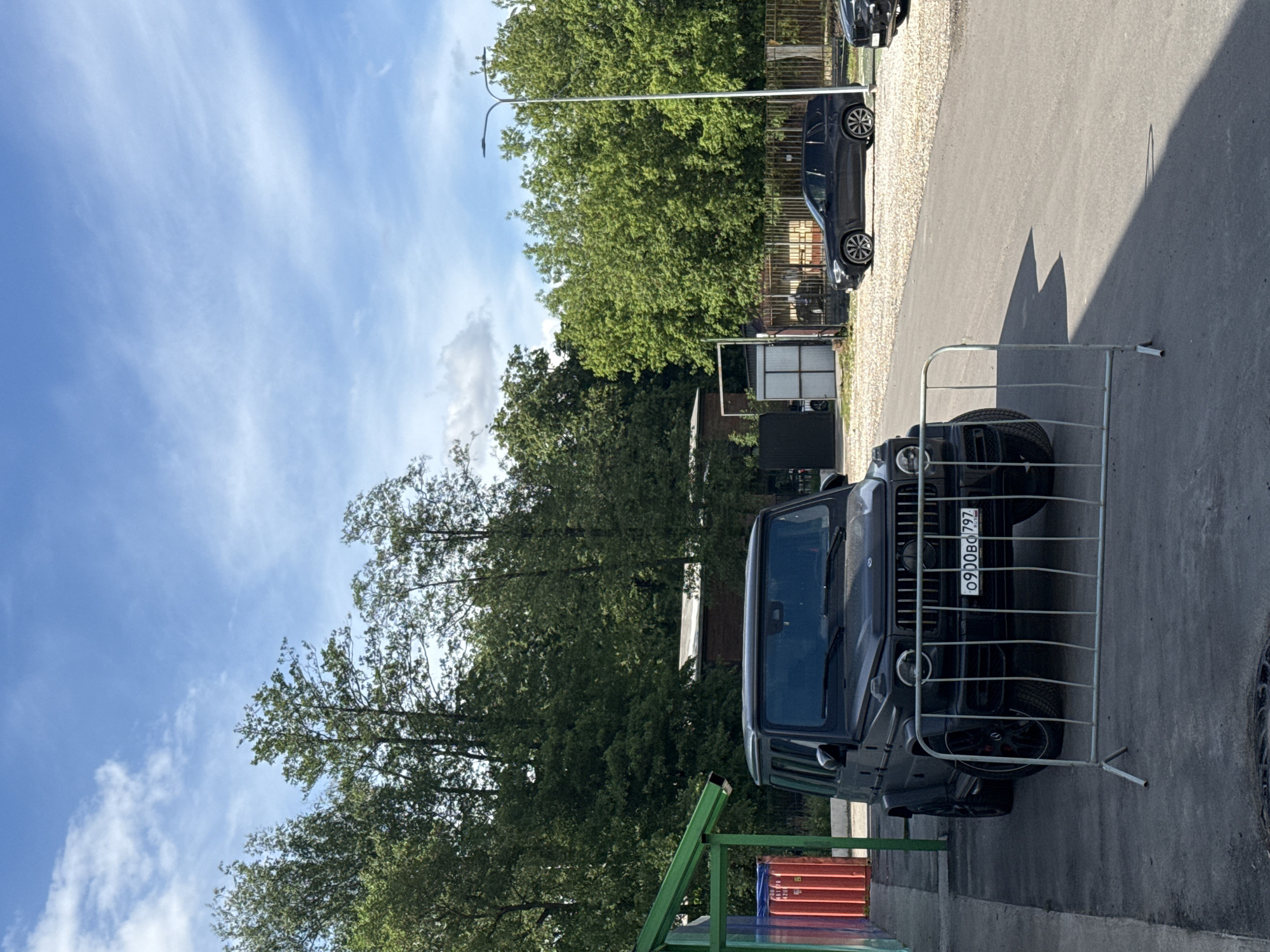 The image depicts a black Mercedes-Benz G-Class parked on an asphalt surface. A metal barrier is positioned in front of the vehicle, partially obscuring its front. A license plate with the registration "O900BO797" is visible. The background includes trees, a light pole, and other parked cars. The scene is located in the city of Moscow, Russia.