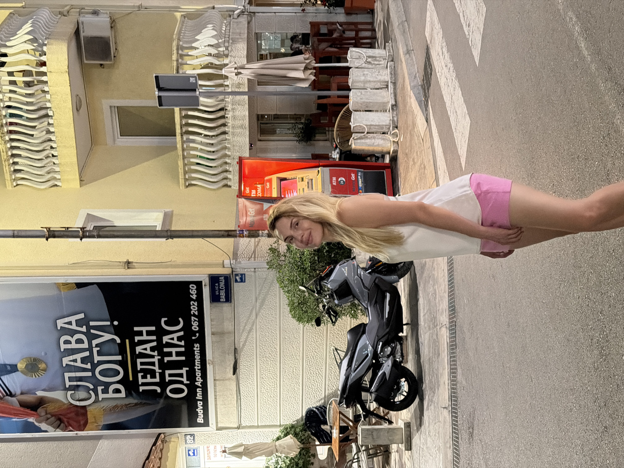 The image shows a woman standing on a street in Budva, Montenegro. She is wearing a white tank top and pink shorts. The street is paved and there is a pedestrian crossing in front of her. Behind her, there is a scooter parked on the sidewalk, a billboard advertising "Budva Inn Apartments" and an ATM machine. The buildings in the background are typical of Budva's old town, with yellow facades and white balconies. The lighting suggests it is evening or night.
