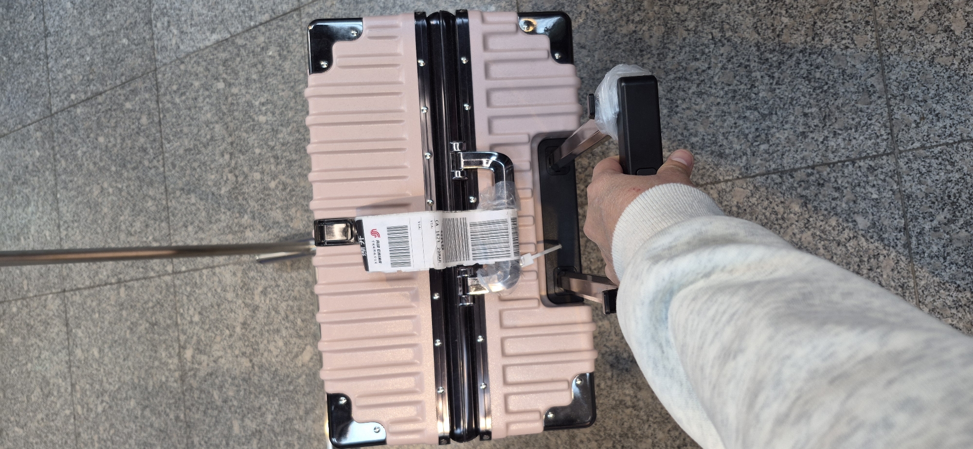 The image shows a person's hand and arm, wearing a light-colored sweater, holding the retracted handle of a light pink, hard-shelled suitcase. The suitcase has a ribbed texture and is adorned with metallic corner protectors and latches. A white baggage tag, featuring a barcode and text that includes "AIR CHINA" and "CA 1629 2DMAF", is attached to one of the suitcase's latches. The suitcase is placed on a gray, speckled floor, likely made of granite tiles, with visible grout lines. In the background, a metallic railing or pole runs horizontally across the frame. The scene appears to be indoors, possibly in an airport or a similar travel terminal, given the flooring and the baggage tag. The lighting suggests it is daytime.