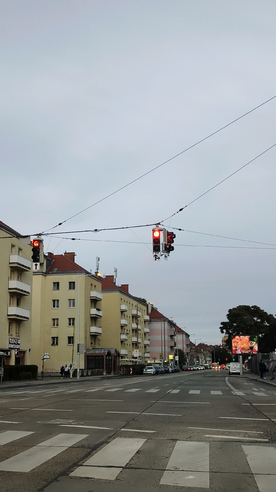 The image shows a street scene in Vienna, Austria, with buildings lining both sides of the road. There are traffic lights overhead, all showing red. Pedestrian crossings are visible on the road. Cars are parked along the side of the road, and some are in motion. A large digital billboard is visible on the right side of the image. The sky is overcast.