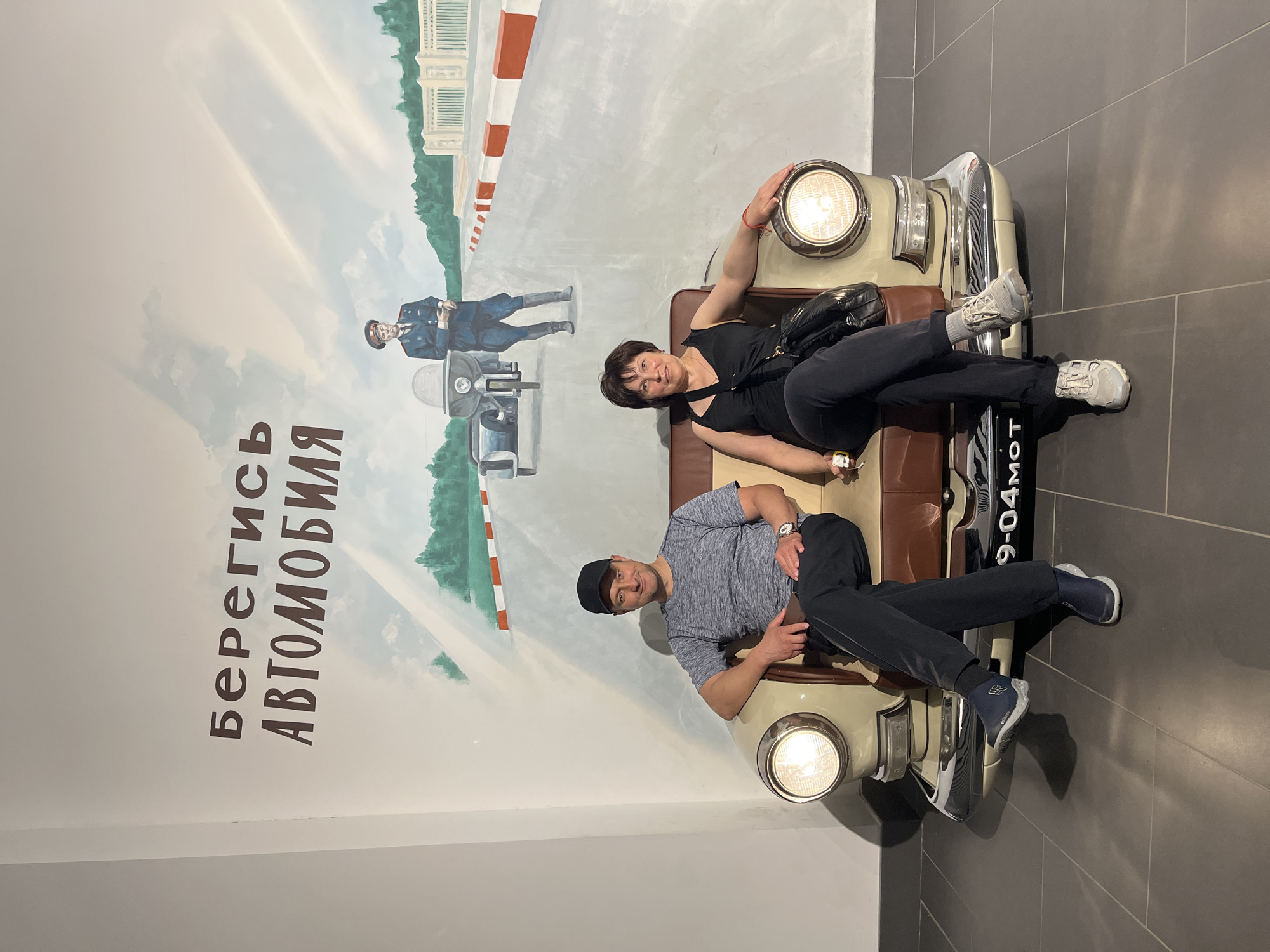 In the Verkhnyaya Pyshma, Russia, the image depicts a man and a woman posing on a prop car. The car, a vintage model with a beige exterior and brown interior, is positioned in front of a mural. The mural features a street scene with a white and orange checkered barrier, a building, and trees. A figure in a blue uniform, possibly a police officer, stands near a motorcycle in the mural.
The woman, wearing a black tank top and black pants, reclines on the car's seat, her arm resting on the headlight. She has dark hair and smiles at the camera. She carries a black bag slung across her body. The man, dressed in a grey t-shirt, black pants, and a black baseball cap, sits on the other side of the car, looking towards the camera with a smile. He wears a watch on his left wrist. Both are wearing athletic shoes.
The backdrop is a white wall with the mural. On the left side of the mural, text is visible in large, dark brown Cyrillic letters: "Берегись АВТОМОБИЛЯ" which translates to "Beware of the car".
The scene appears to be indoors, possibly at a museum or exhibition, given the presence of the prop car and the stylized mural. The lighting suggests an indoor environment, with no indication of natural light or weather conditions. The overall mood is playful and lighthearted, with both individuals smiling and posing for the photograph.