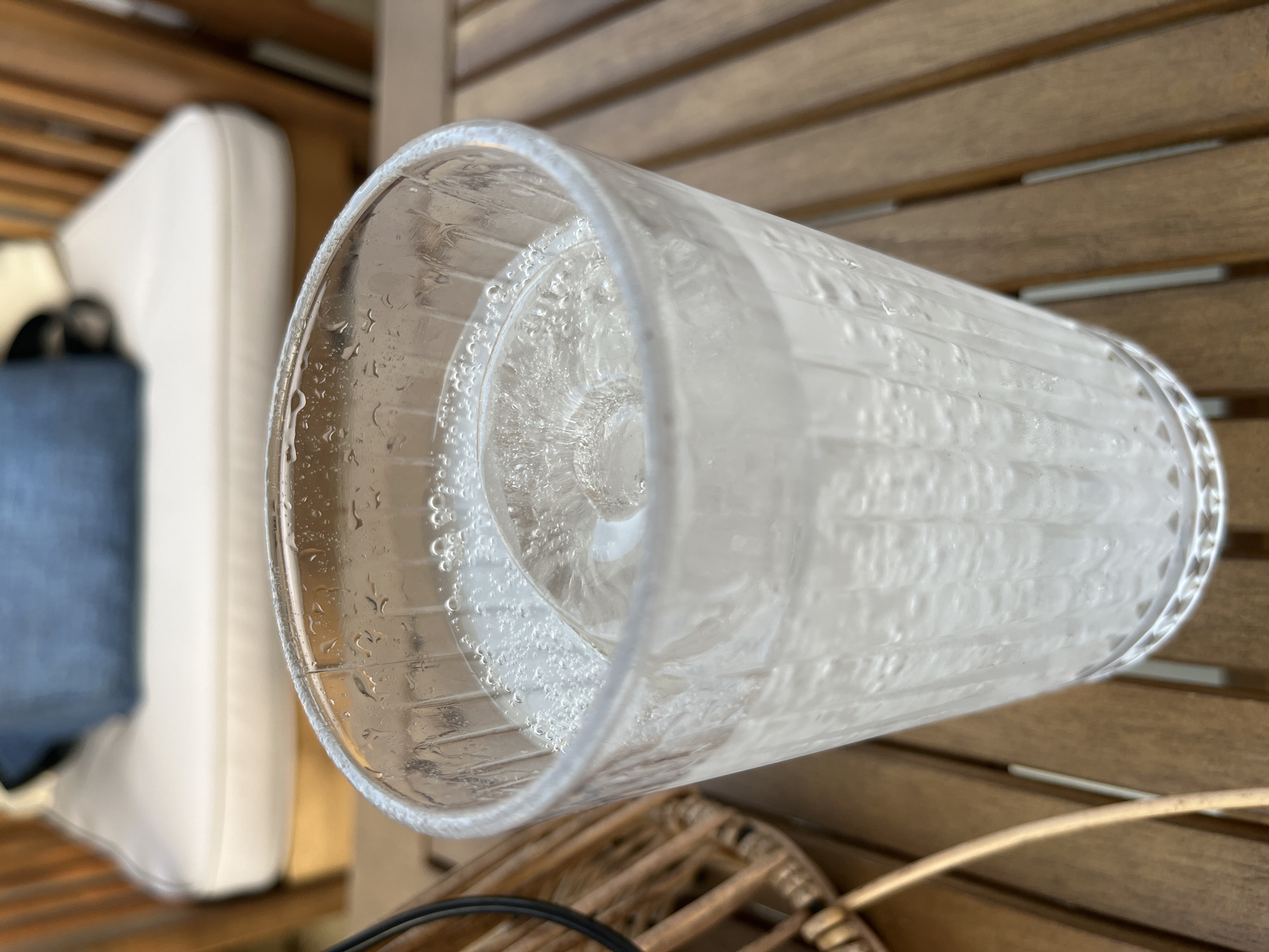 A close-up shot focuses on a clear glass filled with a carbonated beverage and ice. The glass, with its fluted exterior, sits on a wooden slatted table. Water droplets bead on the outside of the glass, suggesting it is cold. In the background, out of focus, is outdoor furniture: a wooden chair with a cushioned seat and a blue pillow. The lighting is natural, casting a soft glow on the scene. The image does not depict any specific location within Nha Trang, Vietnam; it is a generic outdoor setting. There are no people or animals present.