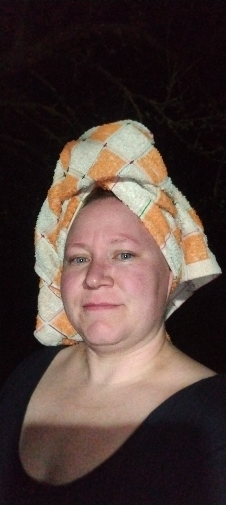 The image shows a woman with a towel wrapped around her head in a turban style. The towel has a checkered pattern of orange and white. She is wearing a dark-colored, low-cut top. The background is dark and indistinct, possibly suggesting an outdoor setting at night. It is impossible to ascertain the location as Зипуново, Russia from the image alone.
