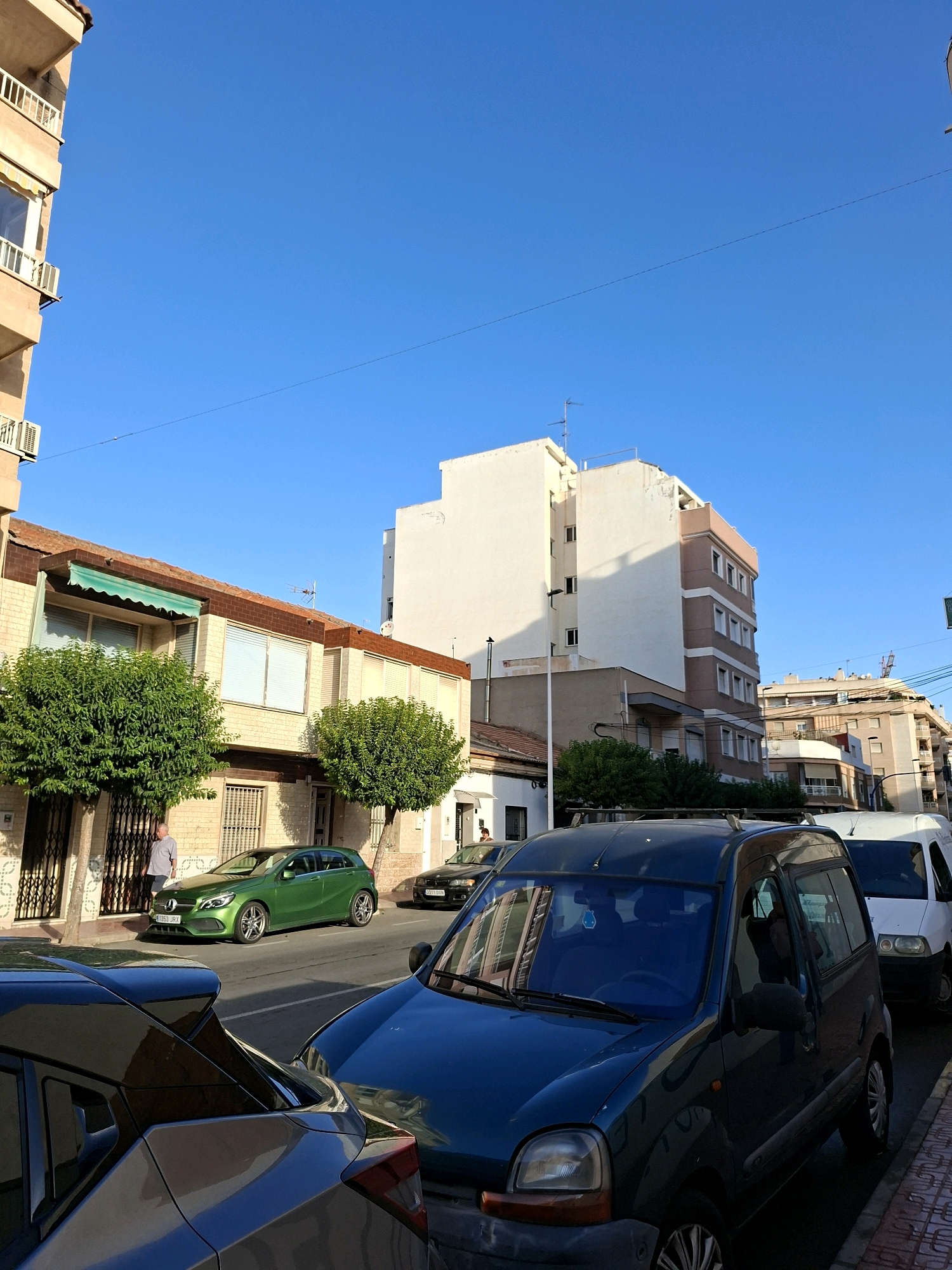 The image captures a street scene in Torrevieja, Spain, under a clear blue sky. In the foreground, a dark blue minivan is parked, partially obscuring a gray car behind it. To the right, a white van is parked on the side of the road. Further down the street, a bright green hatchback is parked, and a man in a grey shirt stands near a building with a green awning. Several multi-story buildings line the street, some with balconies and windows. Trees are planted along the sidewalk. The scene appears to be a residential area with a mix of older and more modern architecture.