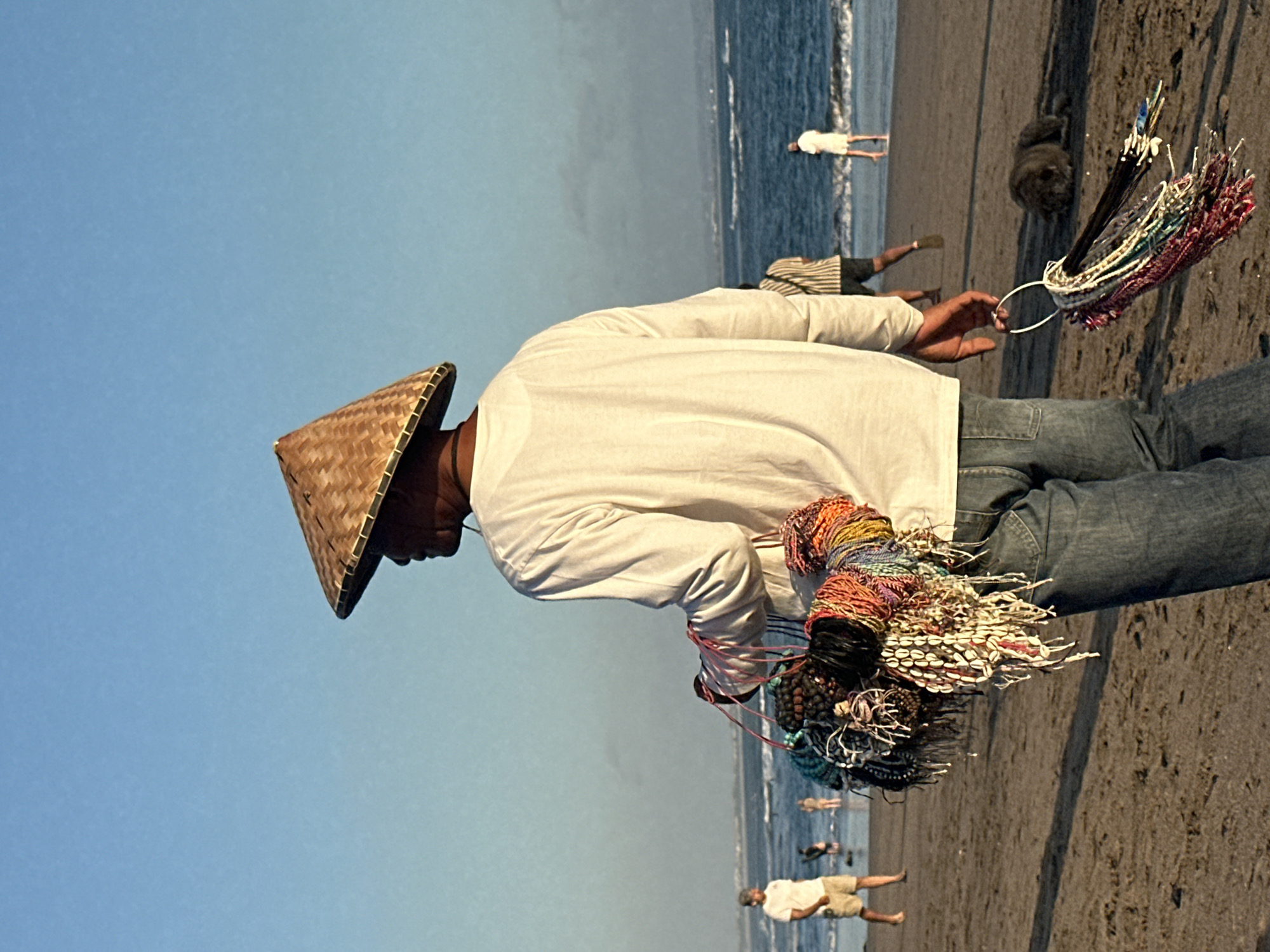 A person wearing a conical straw hat and a white long-sleeved shirt stands on a beach, holding a bunch of colorful bracelets. The person is wearing blue jeans. The bracelets appear to be made of beads, shells, and other decorative materials. In the background, the ocean is visible with waves rolling onto the shore. Several other people are in the distance on the beach and in the water. The sky is a clear blue, suggesting a sunny day. The scene appears to be set in North Kuta, Indonesia. The lighting suggests it is either early morning or late afternoon. The overall mood is calm and perhaps a bit touristy. There is no visible text in the image.