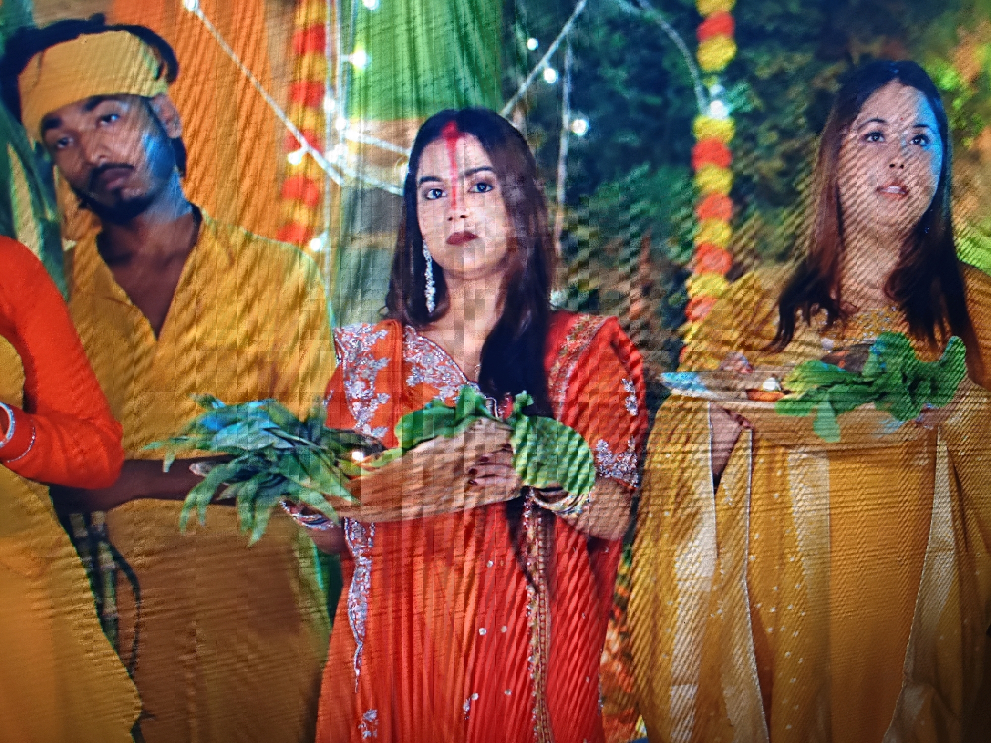 In Garhwa, India, three individuals are depicted in what appears to be a ceremonial or celebratory context, set against a backdrop decorated with colorful floral garlands and string lights. On the left, a man with a yellow bandana and a yellow shirt is visible from the chest up, his face turned towards the camera. In the center, a woman in an orange, ornate dress with silver embroidery and a red bindi on her forehead holds a bundle of green leaves and what seems to be a small lamp or diyā in her hands. To her right, another woman, wearing a golden-yellow garment with white polka dots, also holds a tray with green leaves and a small lamp. The lighting suggests it is evening or night. The overall impression is one of a religious or cultural observance.