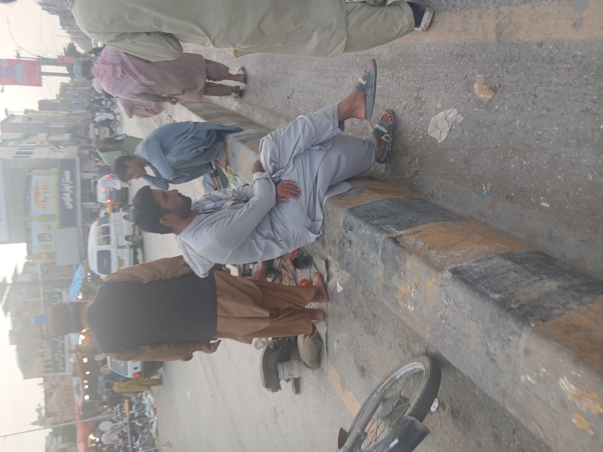The image shows a street scene in Quetta City Tehsil, Pakistan. Several people are visible, including a man lying on a curb, wearing light-colored clothing and Skechers slides. Other individuals are walking and standing nearby, some wearing traditional Pakistani clothing like shalwar kameez and waistcoats. The environment includes buildings, vehicles, motorcycles, and street vendors, indicating a busy urban area. The street appears to be paved and there is some litter on the ground. A bicycle wheel is visible in the foreground. The image appears to have been taken during the day.