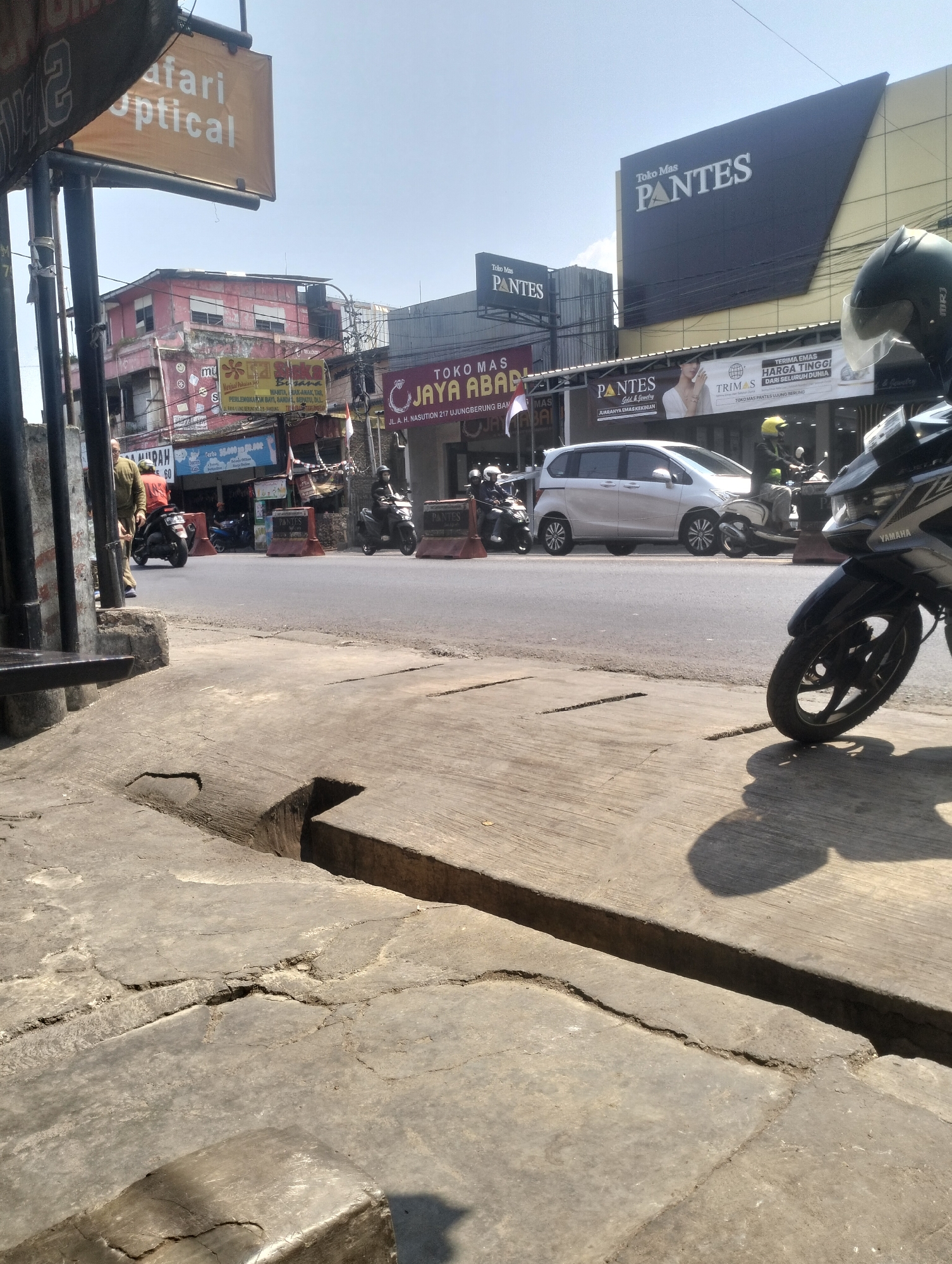 The image shows a street scene in Kel. Pasir Jati, Indonesia, under a clear sky. In the foreground, a cracked concrete sidewalk is visible, with a dark shadow of a person or object cast on it. The street is paved and appears to be a two-lane road with traffic.
Several vehicles are present, including motorcycles and a white minivan. On the street, two people on motorcycles are visible, one wearing a helmet and the other a yellow helmet. There are also traffic cones or barriers on the road.
Along the side of the street are various businesses with signage. One prominent sign reads "Toko Mas PANTES," indicating a jewelry store. Another sign says "SAFARI OPTICAL." Other signs in Indonesian suggest commercial establishments, some with prices listed.
Buildings of varying heights and architectural styles line the street. Some are painted in bright colors like pink, while others have a more subdued appearance. Power lines and poles are visible, typical of an urban environment. The overall impression is a busy, commercial area in Indonesia.