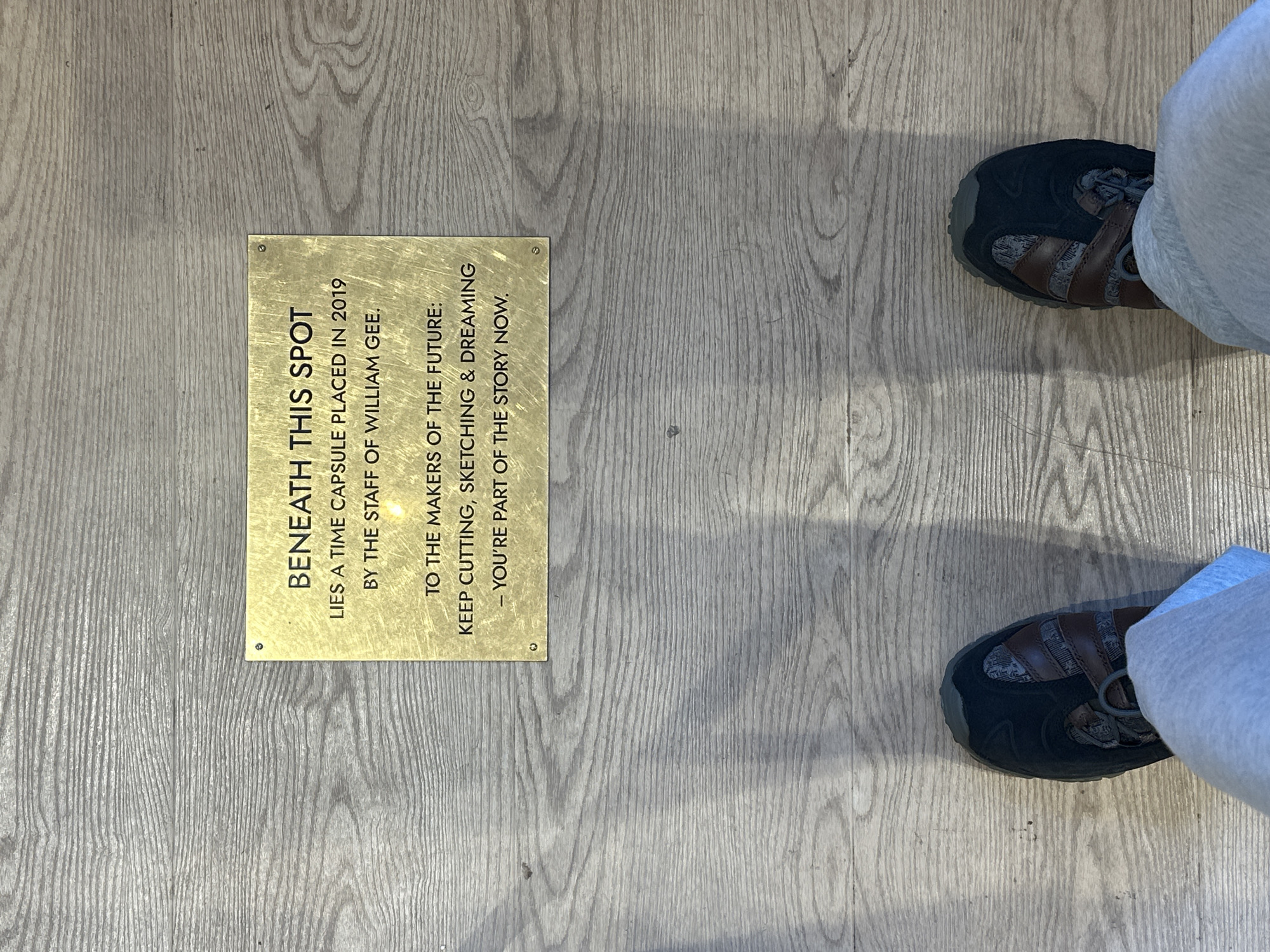 A photograph shows a section of light-colored, simulated wood flooring, on which rests a small, rectangular, brass plaque. The plaque, attached to the floor with screws in each corner, states in black lettering: "BENEATH THIS SPOT LIES A TIME CAPSULE PLACED IN 2019 BY THE STAFF OF WILLIAM GEE. TO THE MAKERS OF THE FUTURE: KEEP CUTTING, SKETCHING & DREAMING - YOU'RE PART OF THE STORY NOW."
To the right of the plaque, two shoes (black with brown accents) and the bottoms of legs wearing light gray sweatpants are visible. The perspective is directly overhead. The location is assumed to be a branch of William Gee, a long-standing shop in Shoreditch, London, which caters to the fashion and design industries.