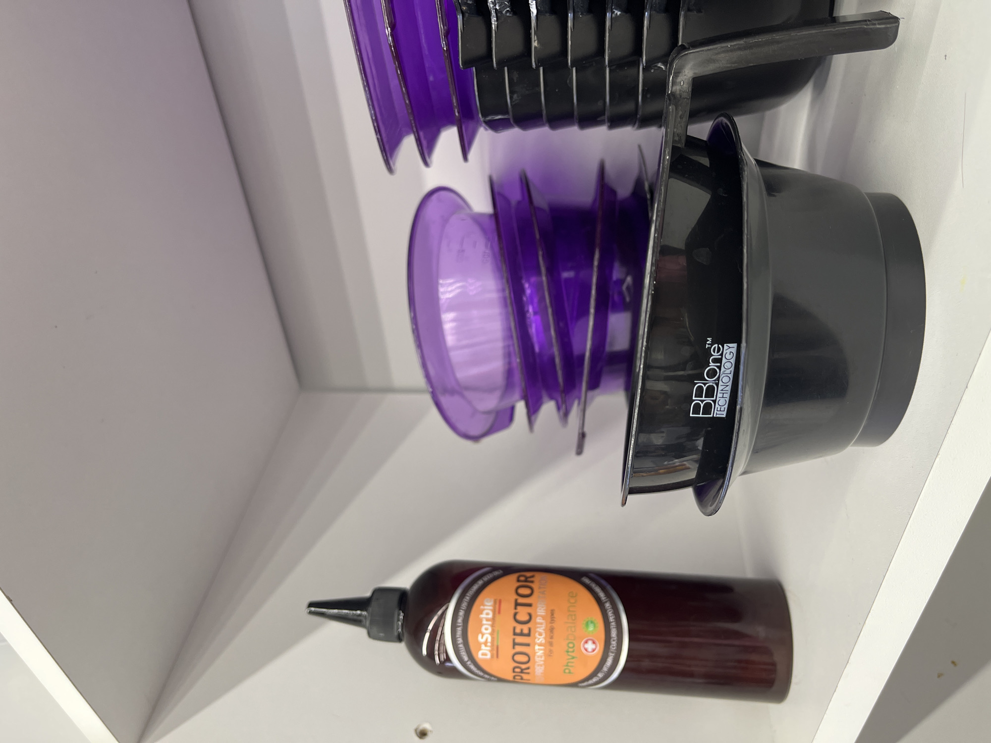 The image depicts a shelf inside a white cabinet. On the shelf are a stack of purple and black hair dye bowls and a bottle of Dr. Sorbie Protector for scalp irritation. The bottle is brown and has a black nozzle.
