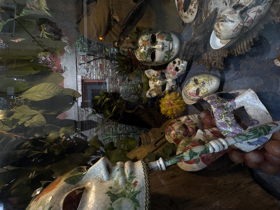 The image depicts a shop window display in Venice, Italy, featuring a collection of decorative Venetian masks. Approximately ten masks are visible, primarily white with a cracked, aged finish, adorned with painted floral patterns in green, red, purple, and blue hues. A prominent half-mask with a gold chain trim and a small gem is positioned on the far left, mounted on a decorated handle. Other masks include a stylized cat face and various full and partial face designs. A single yellow artificial flower and a cluster of reddish-brown, grape-like objects are arranged among the masks, which rest on a dark blue, textured surface alongside pieces of driftwood. The glass window reflects an exterior scene: an aged brick building with a small, barred rectangular window set in a red frame. Dense green foliage from an unseen plant or tree partially obscures the upper portion of the reflected building. No individuals are depicted within the display or the reflection.