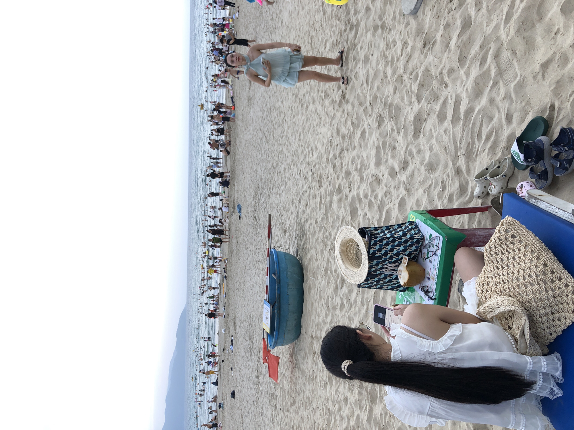The image shows a crowded beach in Da Nang, Vietnam. In the foreground, a woman with long black hair is sitting on a blue mat, looking at her phone. In front of her is a small green and red table with a straw hat, a patterned bag, glasses, and a coconut with a straw. To her right, another woman in a light blue dress is standing and looking at her phone. The background is filled with people swimming and sunbathing in the ocean and on the sand. A blue boat with red flags is on the beach near the water. The sky is overcast, and mountains are visible in the distance. Several pairs of sandals and flip-flops are scattered on the sand near the foreground.
