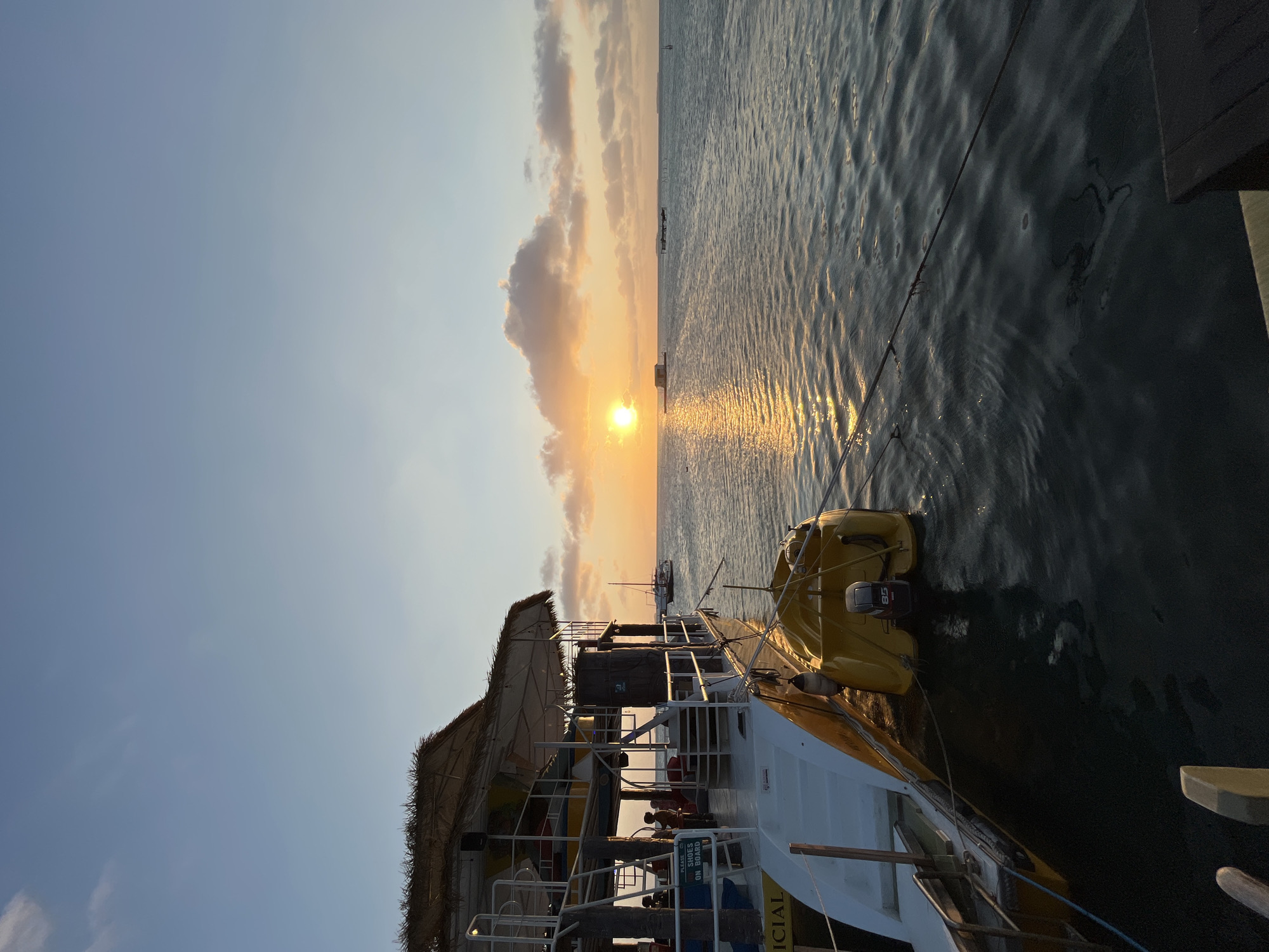 The image depicts a sunset scene at a dock in Karimunjawa, Indonesia. A moored boat with a thatched roof structure is prominently featured in the foreground on the left. The boat has a white and yellow hull and a smaller yellow dinghy is attached to its side. Several people are visible on the boat's deck. The sun is setting over the ocean on the right side of the frame, casting a golden reflection on the water. In the distance, there are other boats and structures on the water, as well as a hazy horizon. The sky is a gradient of blue and orange, with scattered clouds.