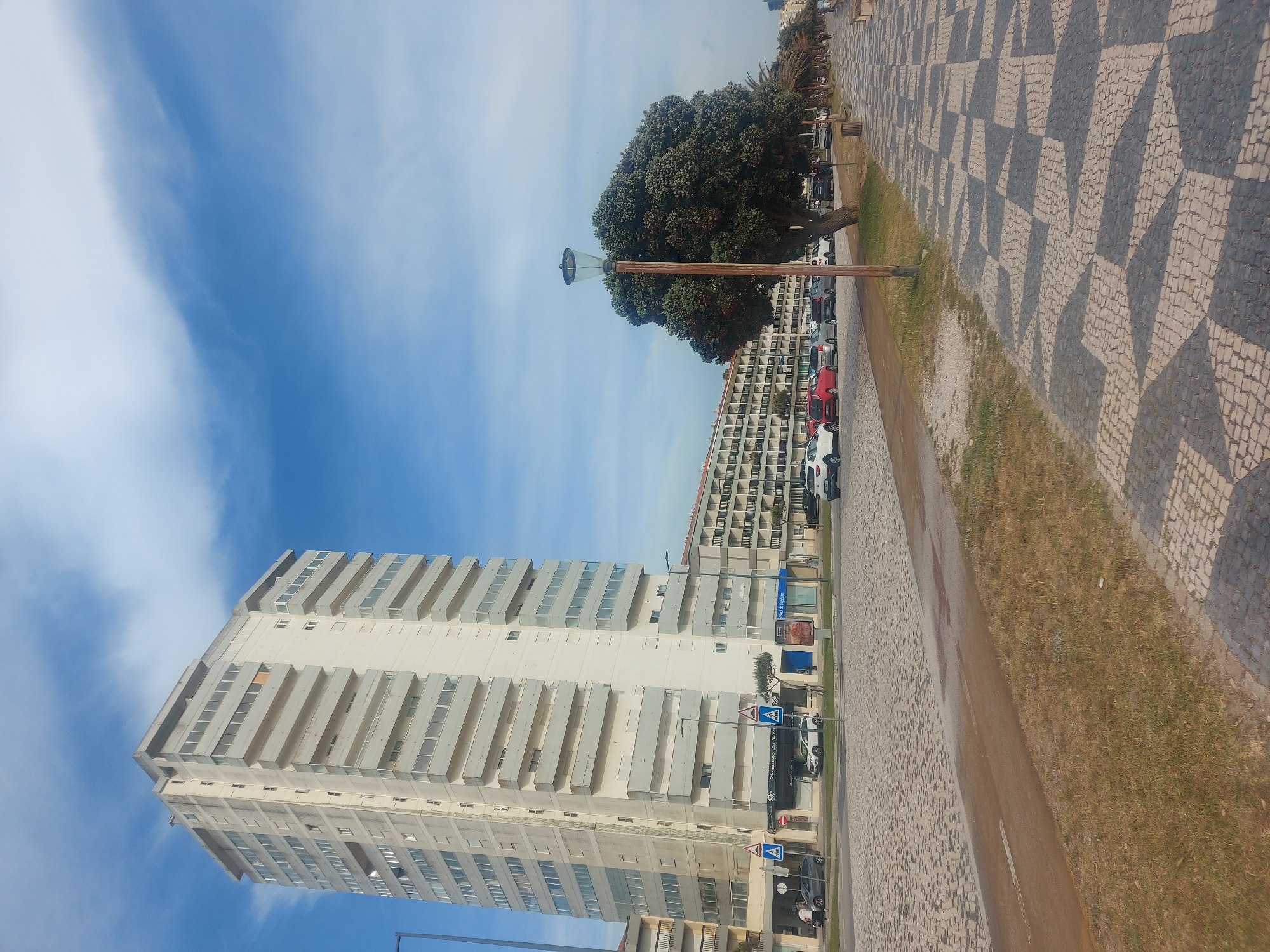 The image captures a bright daytime scene in Figueira da Foz, Portugal, under a clear blue sky with sparse white, wispy clouds. The foreground and right side of the frame are dominated by a distinctive patterned mosaic pavement, characteristic of "calçada portuguesa," featuring interlocking geometric shapes in light and dark grey tones. This paving extends into the background, bordering a strip of dry, light brown grass, which in turn gives way to a road with parked cars.
On the left, a tall, slender, light-colored multi-story apartment building stands prominently, characterized by numerous horizontal balconies creating a stepped facade. In the mid-ground, a large, dense tree with a rounded green canopy partially obscures a modern streetlamp, which has a grey bell-shaped head and a brown pole. Behind the tree and across the road, another multi-story building with numerous balconies is visible, and more cars are parked along this street.
Several road signs and advertisements are discernible. A blue rectangular sign clearly displays "Gest. de Imóveis" (Real Estate Management). Another blue sign below it is partially obscured but starts with "Guesthouse da R...". Other visible signs include multiple blue square pedestrian crossing signs with white figures, a circular red 'no entry' sign, and a blue circular 'turn right' sign. Small, indistinct figures of people can be seen near the buildings and parked vehicles. The scene presents a typical, sunny urban street environment.