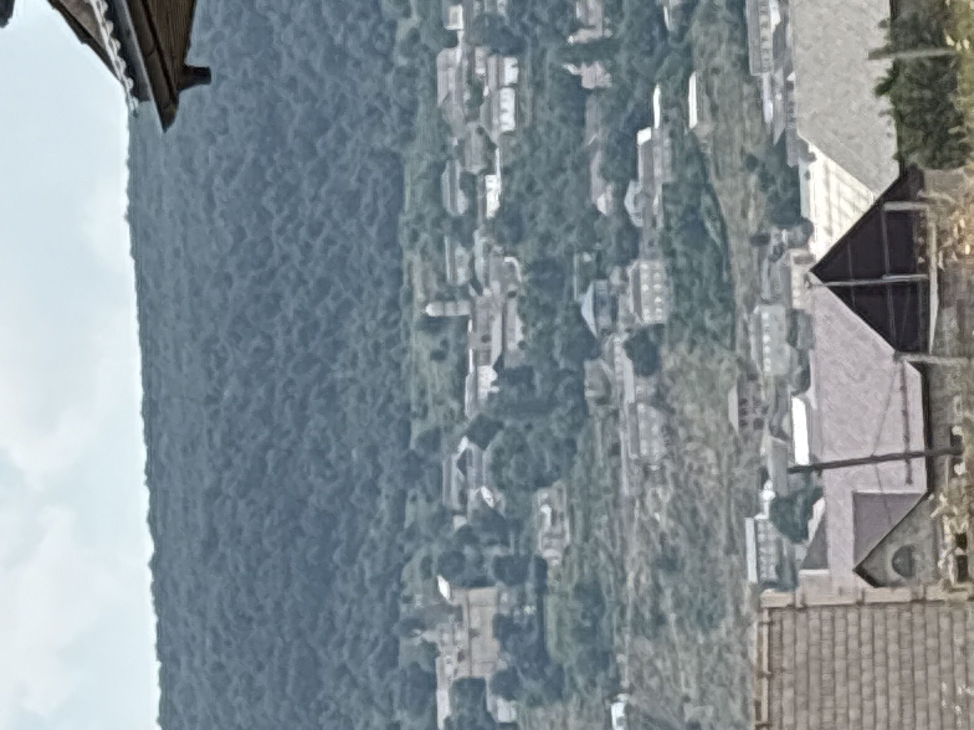 A wide shot captures a hillside village with buildings nestled amongst dense green foliage and trees under a hazy sky. The village, identified as being in Ullubiyaul, Russia, features a mix of architectural styles, from older stone structures to more modern residential buildings with distinct rooflines. In the foreground, the roofs of several structures are visible, including one with a prominent triangular dark roof. A tall, thin pole stands vertically to the right of this structure. The hillside is terraced in some areas, suggesting agricultural activity. The overall impression is one of a rural, somewhat isolated community. No people or specific actions are discernible in the image.