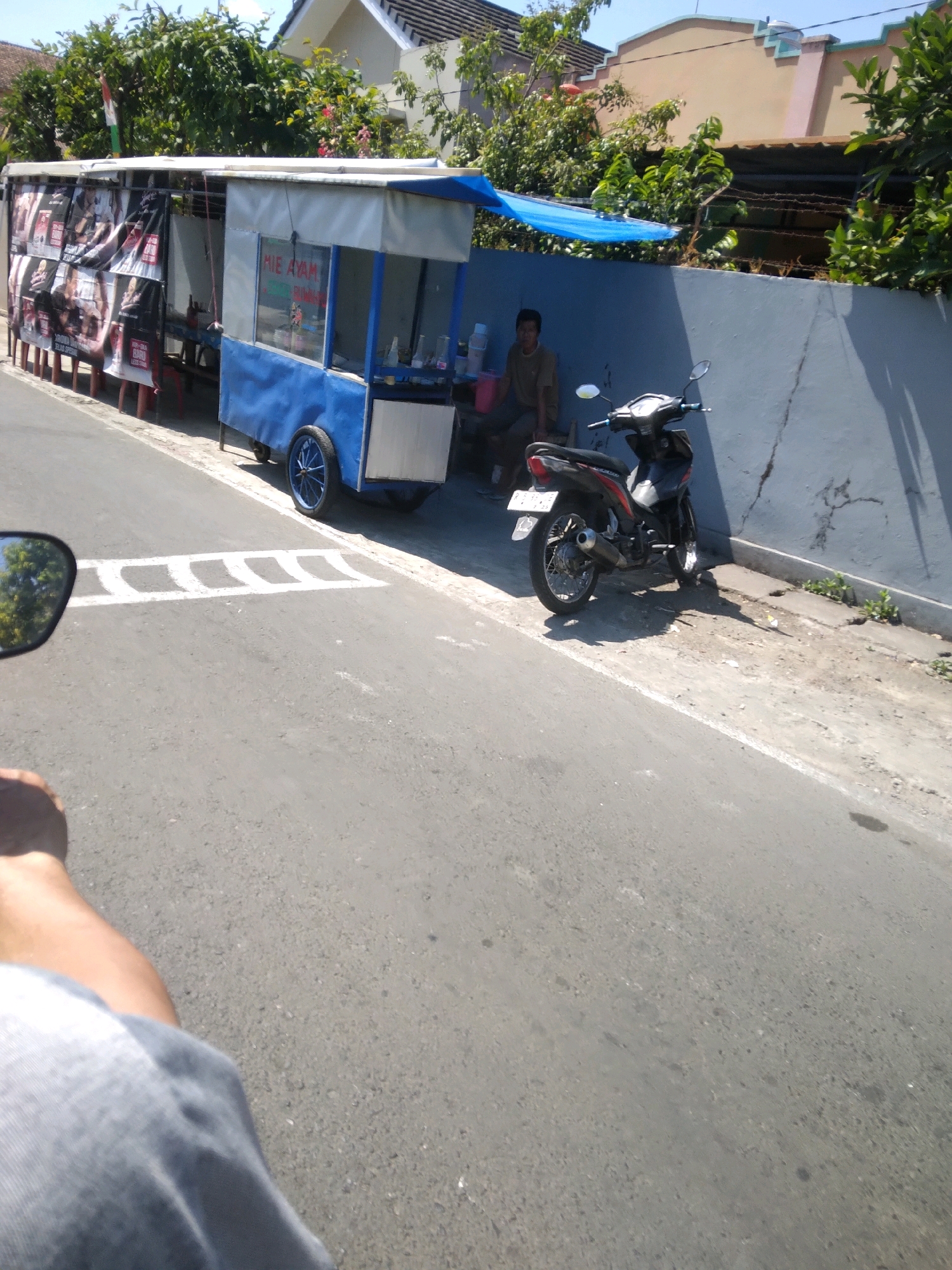 The image depicts a street scene in Purwokerto Wetan, Indonesia, under bright daylight conditions. A food cart selling "MIE AYAM" (chicken noodles) is prominently featured, with its blue and white structure. Posters are displayed on the side of the cart. A man, dressed in a brown shirt, is seated near the cart, appearing to be the vendor. A motorcycle is parked next to the cart, leaning on its stand. The street itself is paved with asphalt, and a white stripe marking a speed bump is visible. In the foreground, a portion of a person's leg and arm, likely the photographer's, are visible, suggesting the photo was taken from a moving vehicle, possibly a motorcycle. The background shows residential buildings with tiled roofs and lush green trees. The overall atmosphere is that of a typical urban setting in Indonesia on a sunny day.