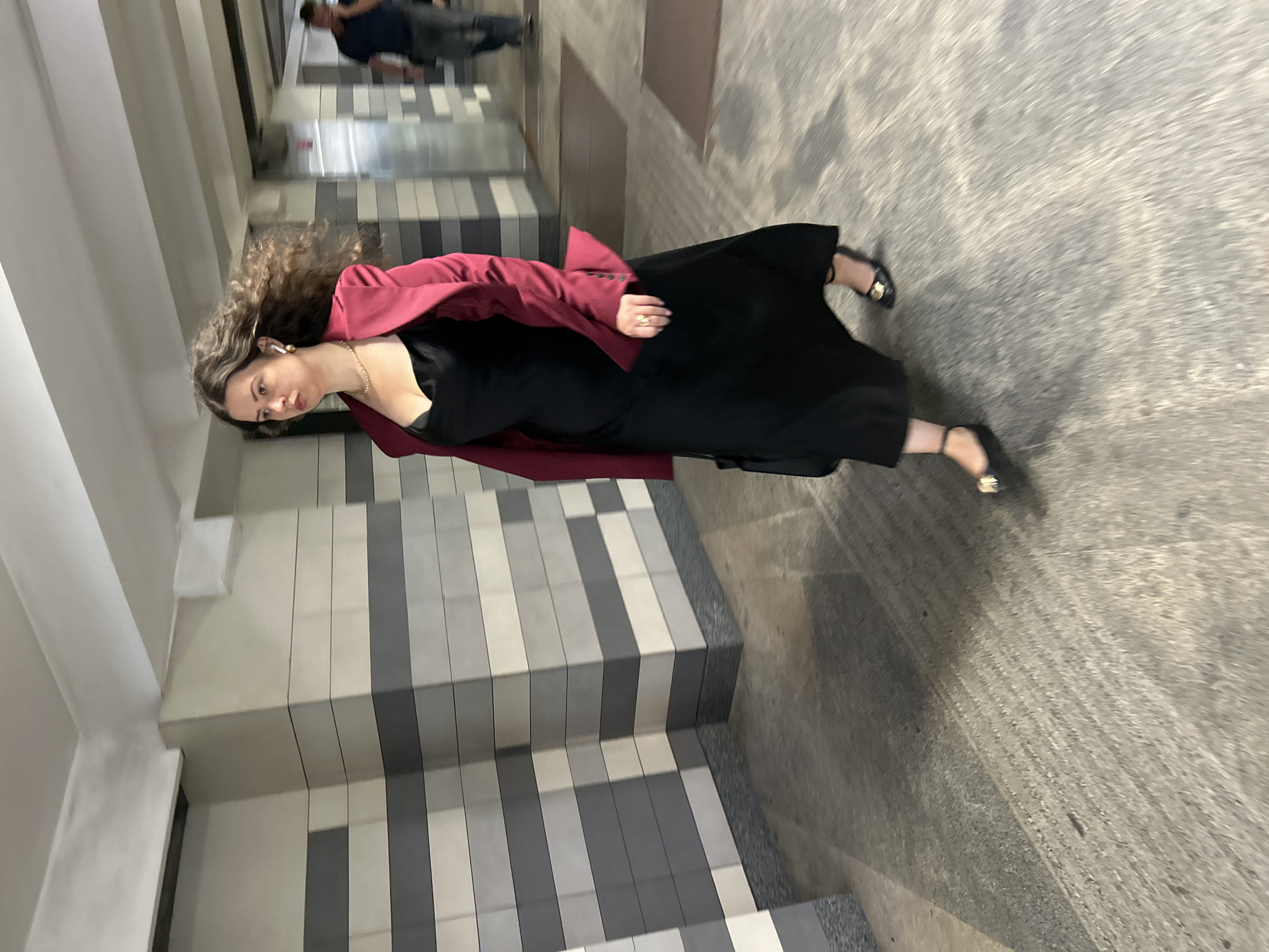 A woman with long, curly brown hair, wearing a black dress and a deep red blazer, walks from left to right in a pedestrian underpass. She has white earbuds in her ears and is wearing a gold necklace and a ring on her right hand. Her black dress is floor-length, and she is wearing black flat shoes with gold accents. She is carrying a black handbag. The underpass has a floor made of textured concrete and walls tiled with horizontal bands of grey and beige. Another person, a man wearing a dark t-shirt and jeans, is visible in the background, walking away from the camera. The lighting is artificial, typical of an urban underpass. This scene is located in Moscow, Russia.